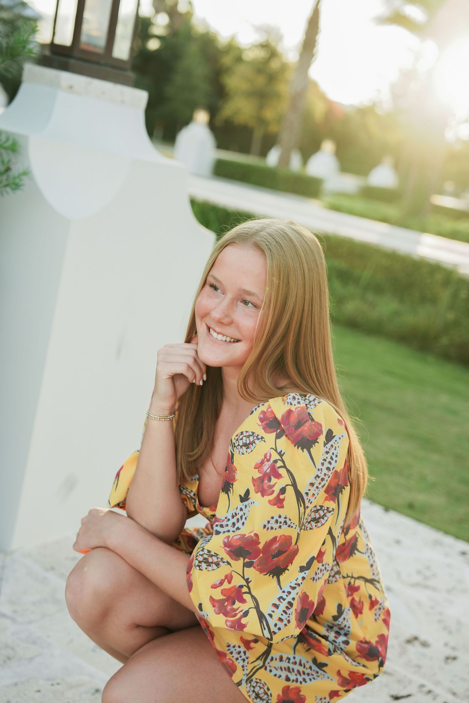 A person wearing a bright yellow floral dress sits on white stone steps in a sunny outdoor setting.