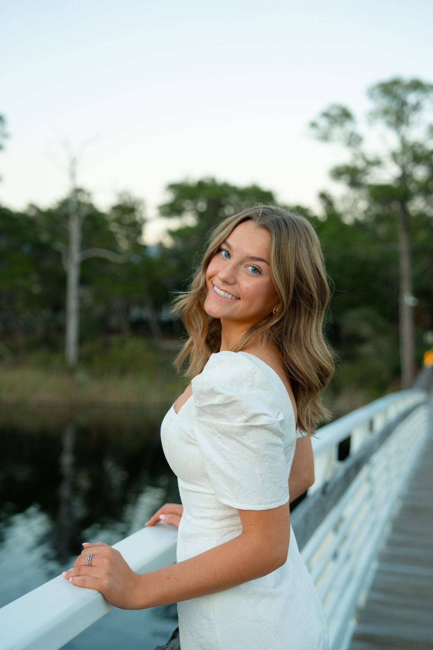 A person in a white top poses joyfully on a wooden pier near water and trees on a bright sunny day.