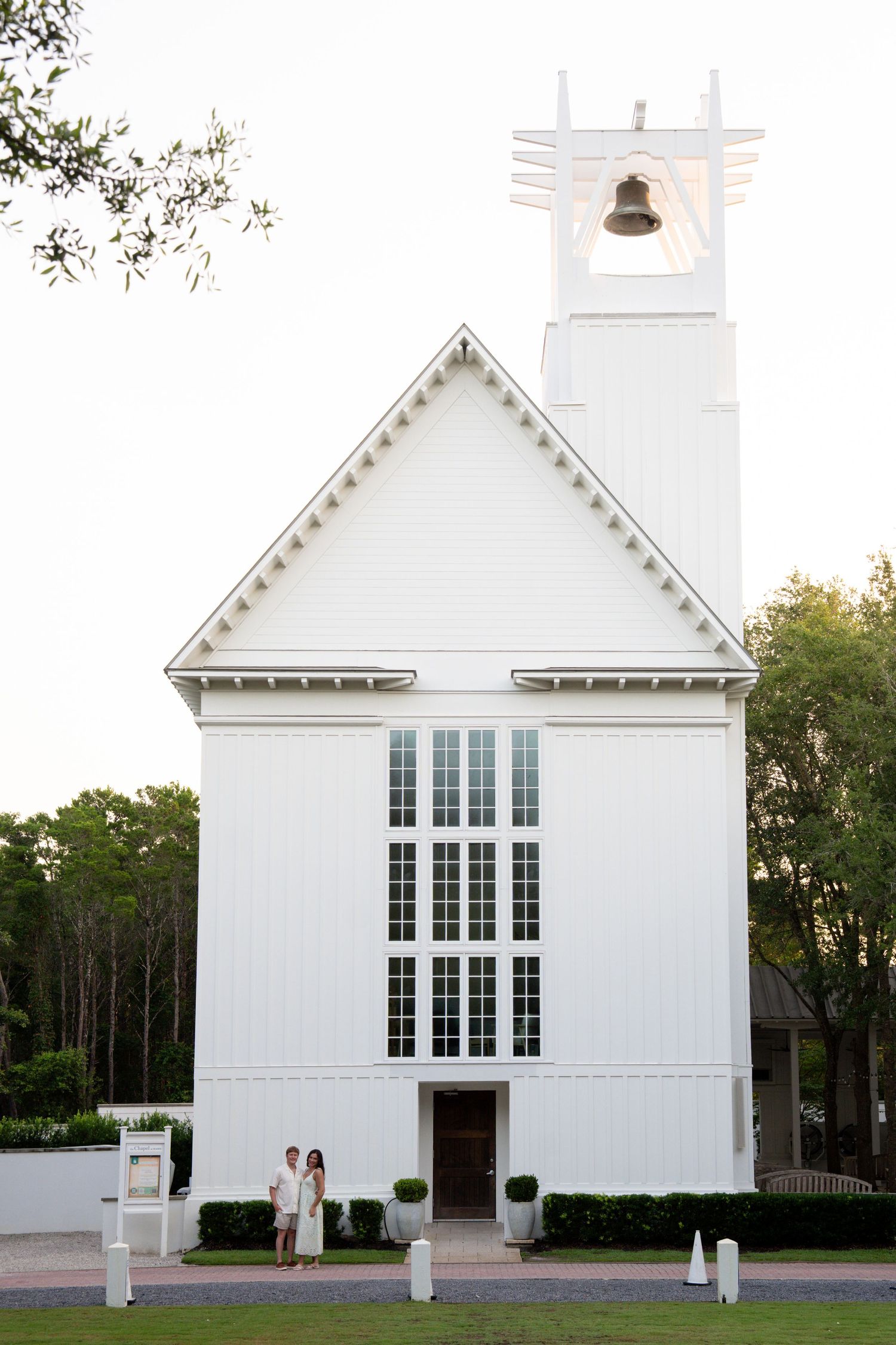 Couple standing in front of a white chapel with bell tower surrounded by lush greenery.