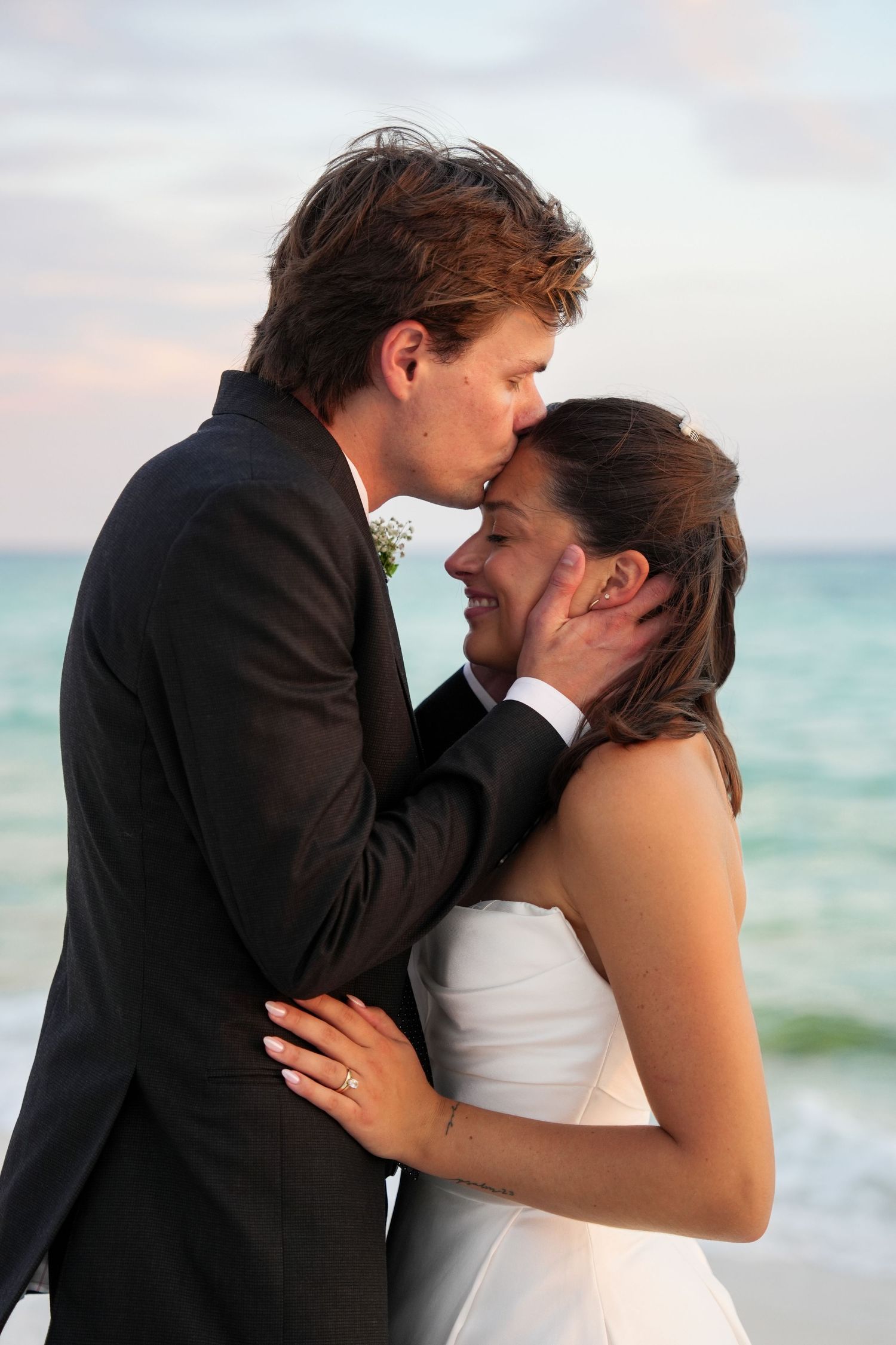 A groom kisses his bride on the forehead during a romantic beach wedding ceremony at sunset.