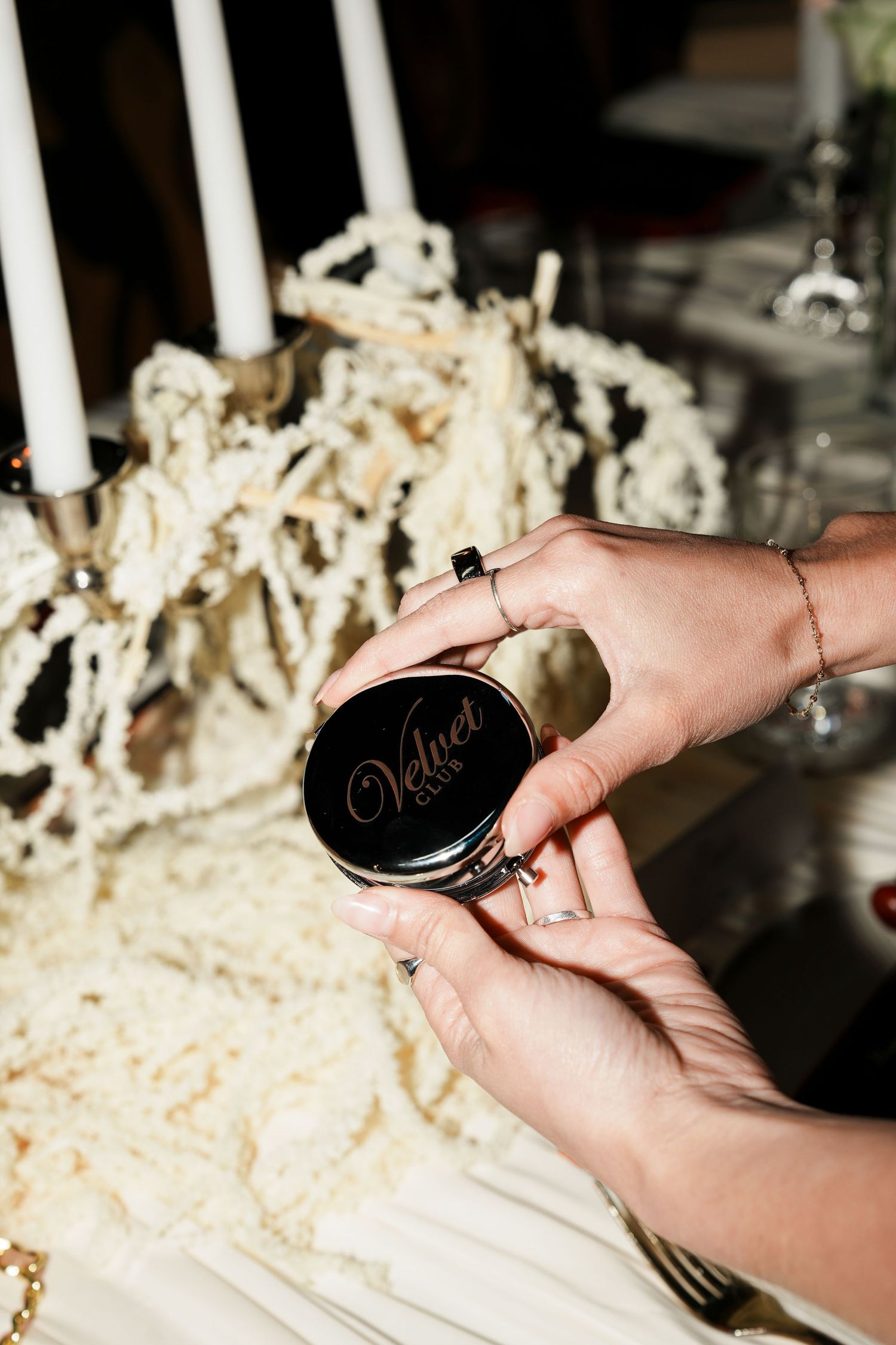Hands holding a black compact mirror engraved with Velvet logo, surrounded by white candles and cream macrame decor.