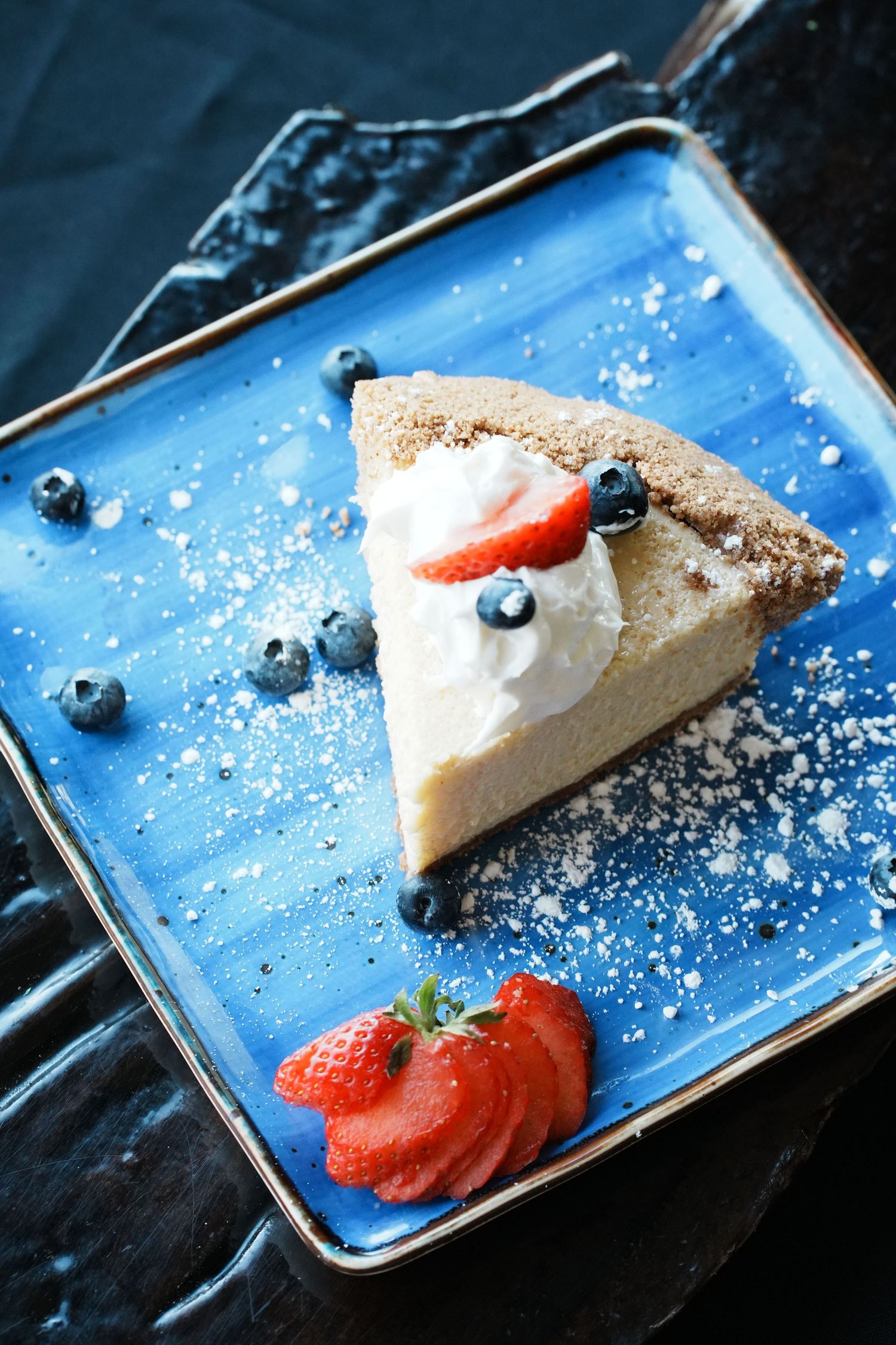 Slice of creamy cheesecake plated with fresh berries, whipped cream and powdered sugar dusting.