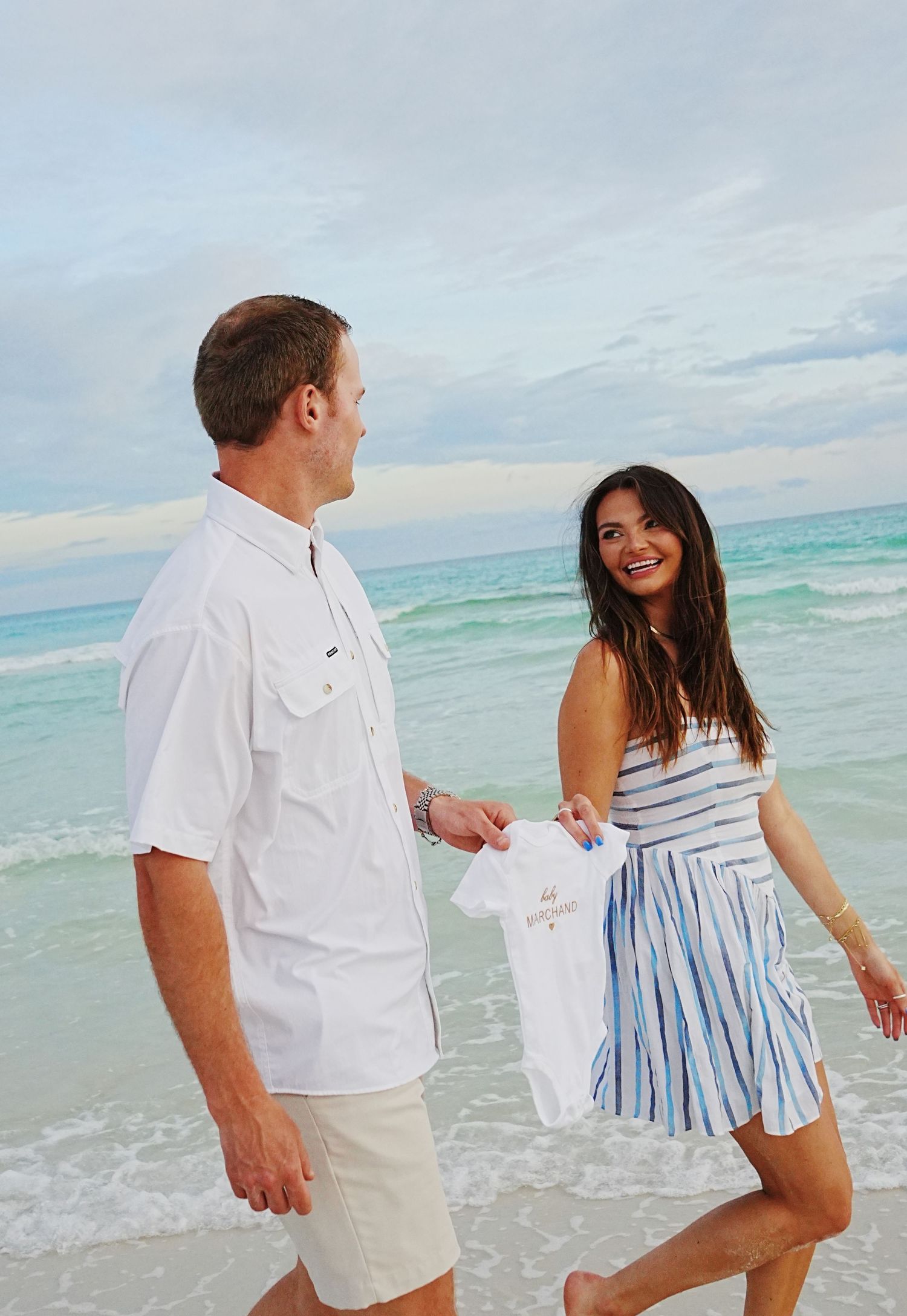A couple enjoying a romantic stroll along a tropical beach with turquoise waters and cloudy skies in the background.