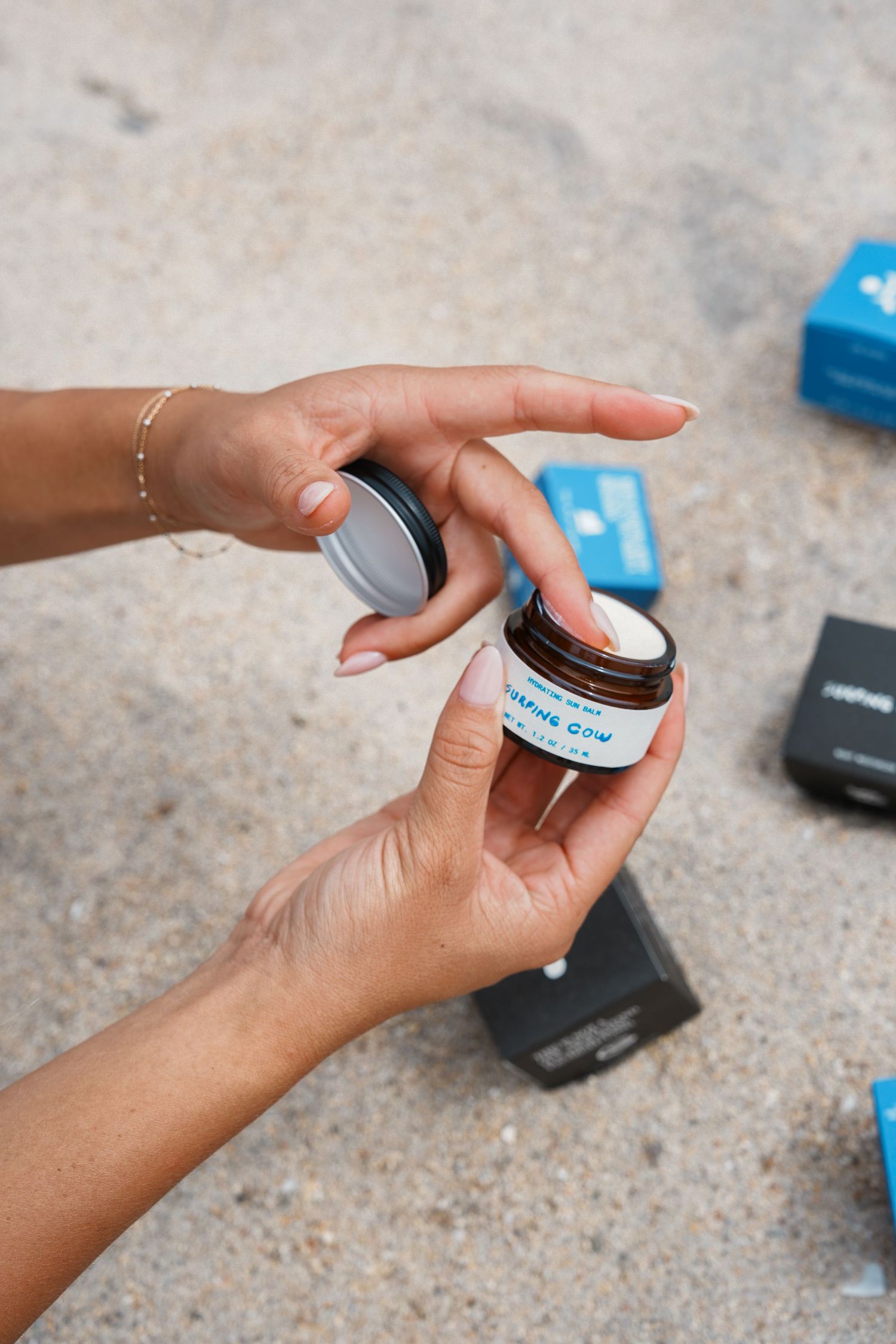 Hands opening a small black cosmetic jar or skincare product container against a textured concrete background.