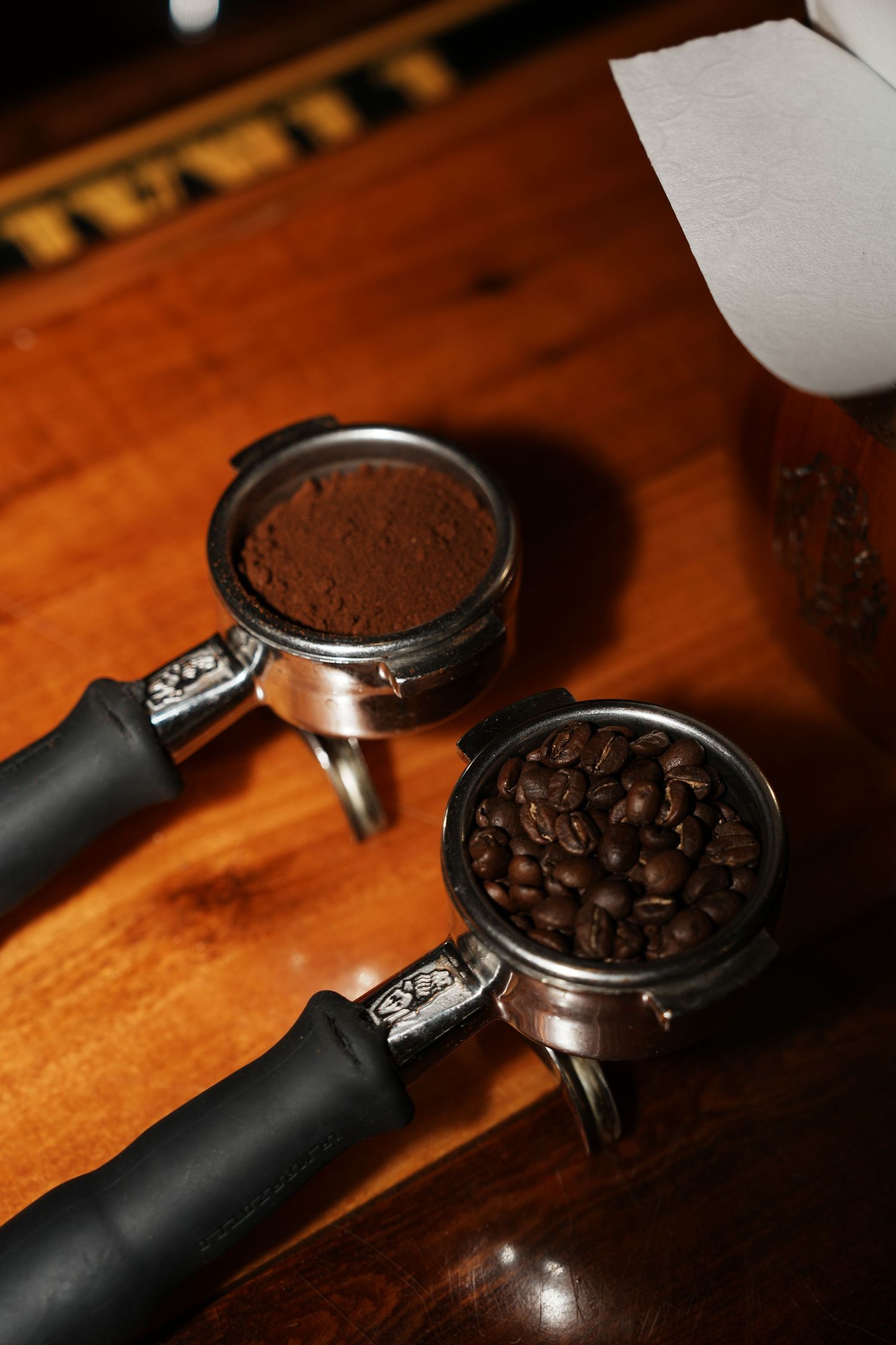 Two espresso portafilters on wooden surface filled with coffee grounds and beans.