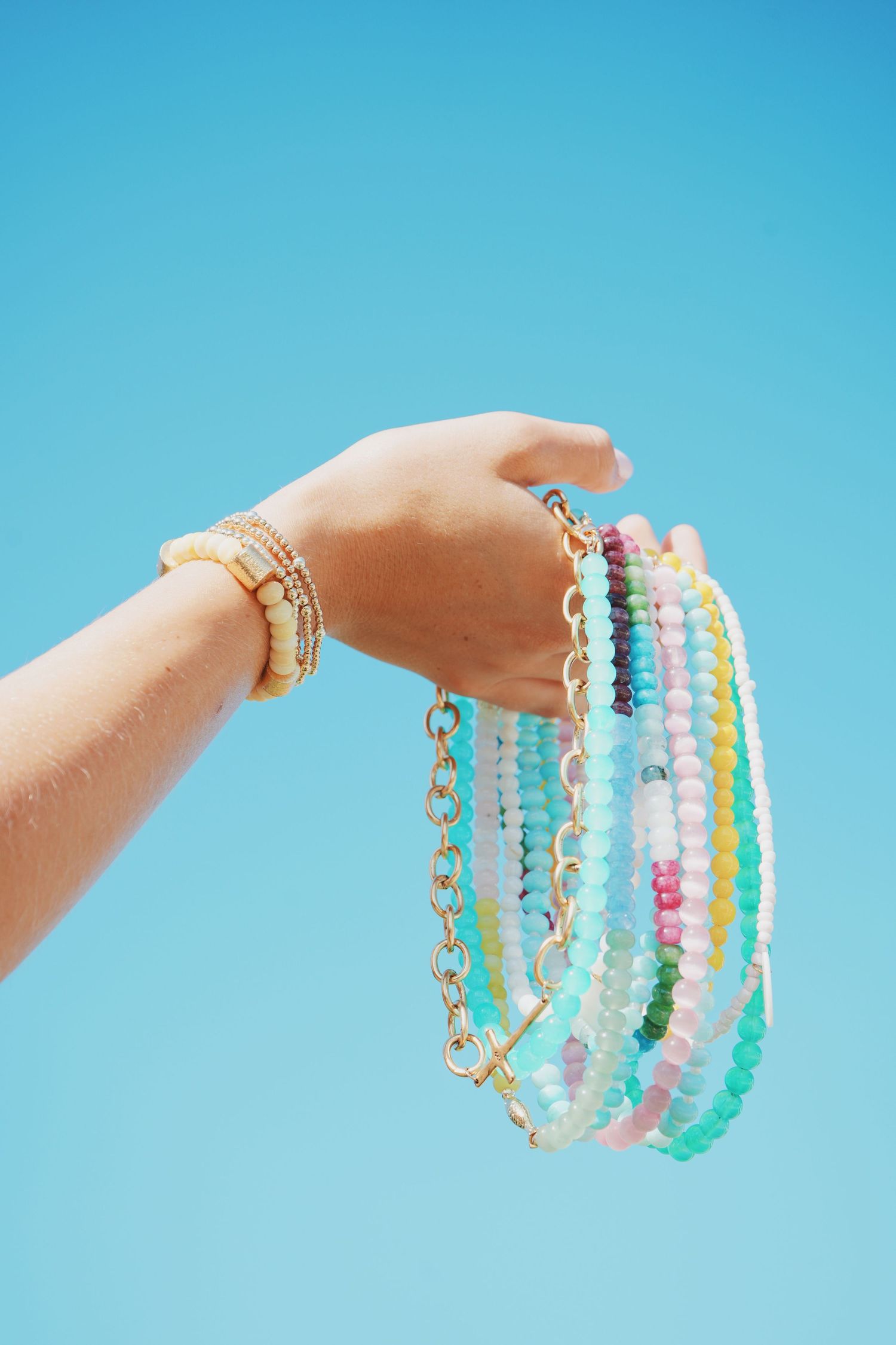 Hand holding colorful beaded necklaces and bracelets against bright blue background.