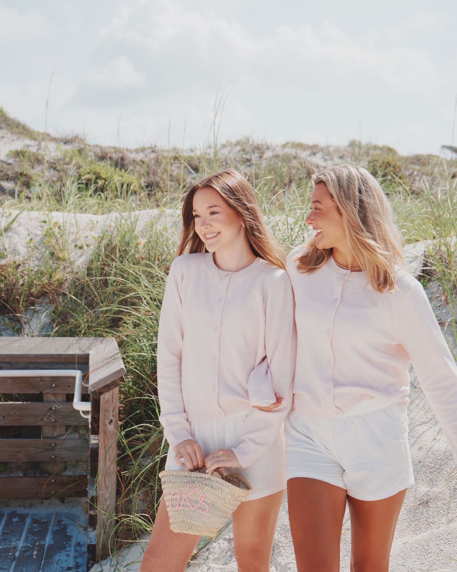 Two friends in matching cream-colored sweatshirts and shorts laugh together on a sandy beach path near tall grass.