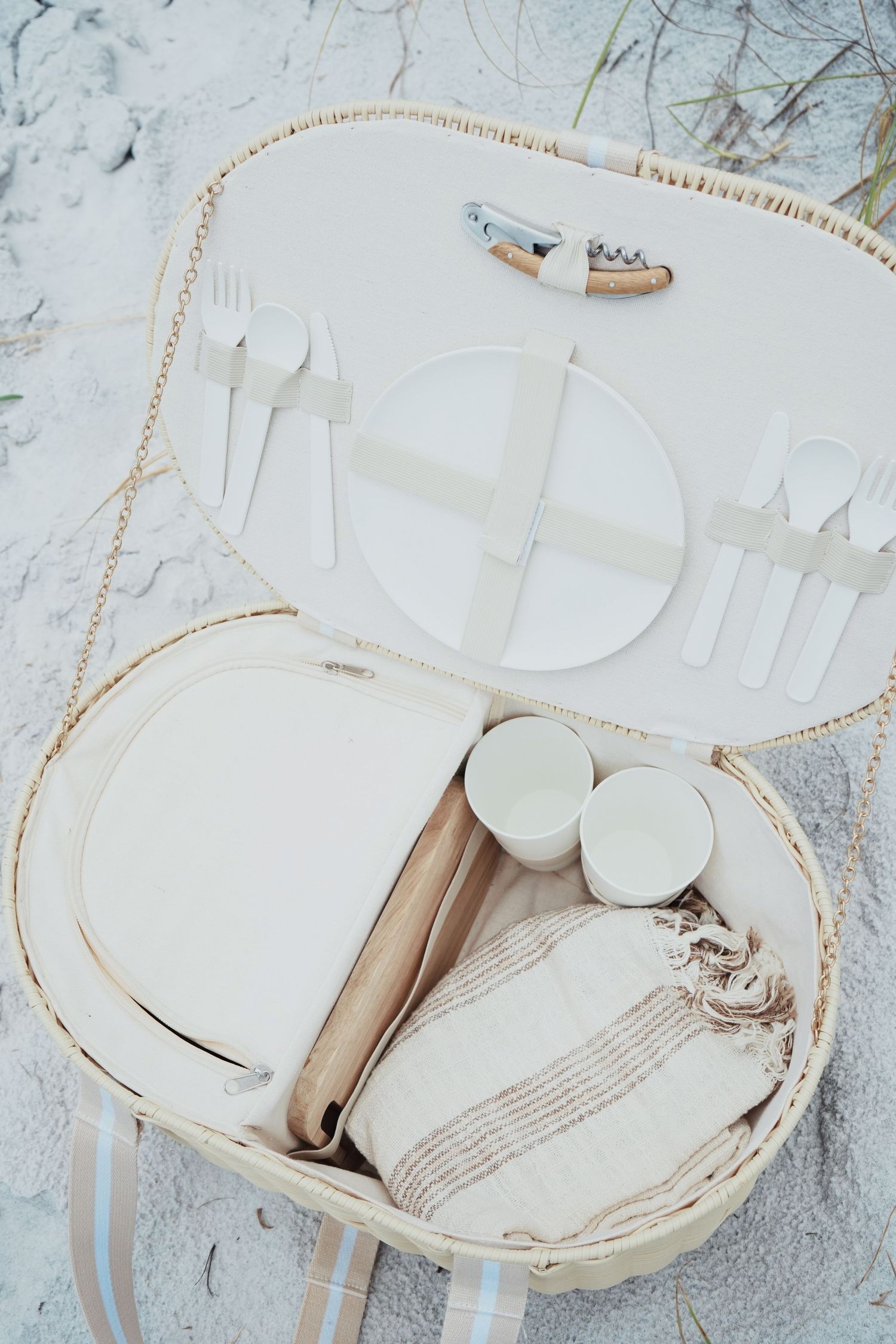 A white vintage picnic basket with plates, utensils, and small cups displayed on a light gray textured surface.