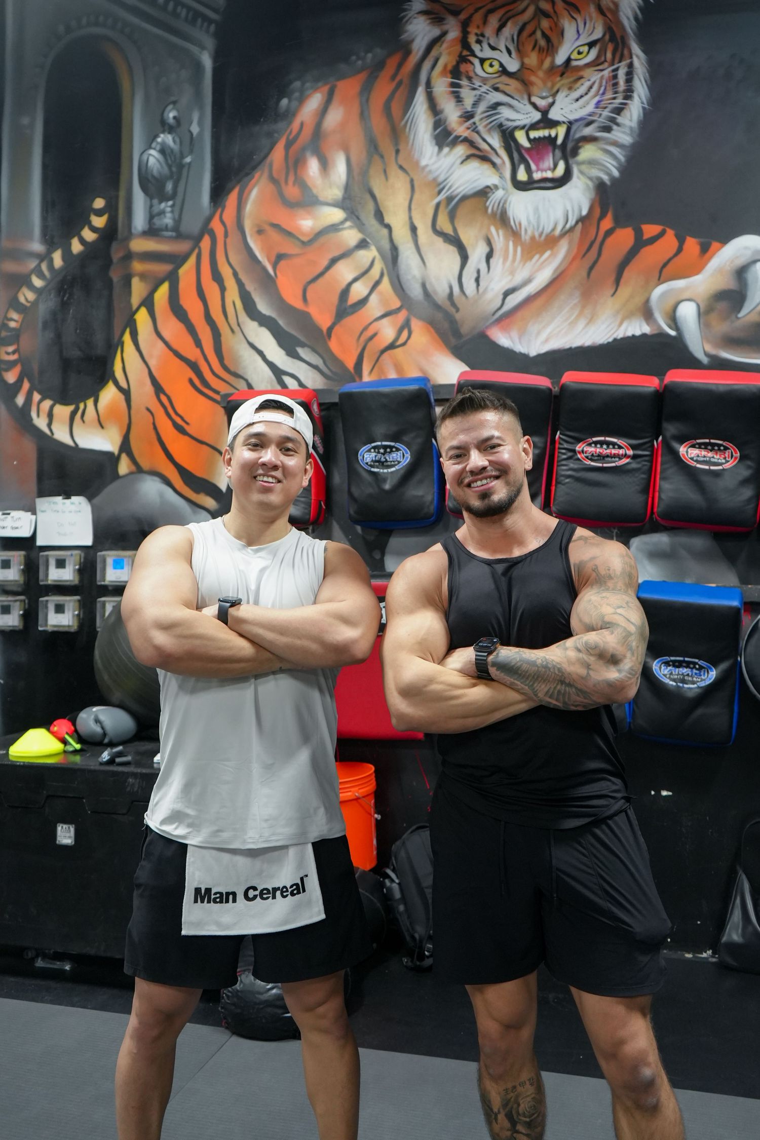 Two fitness trainers pose together in a gym with a large tiger mural painted on the wall behind them.