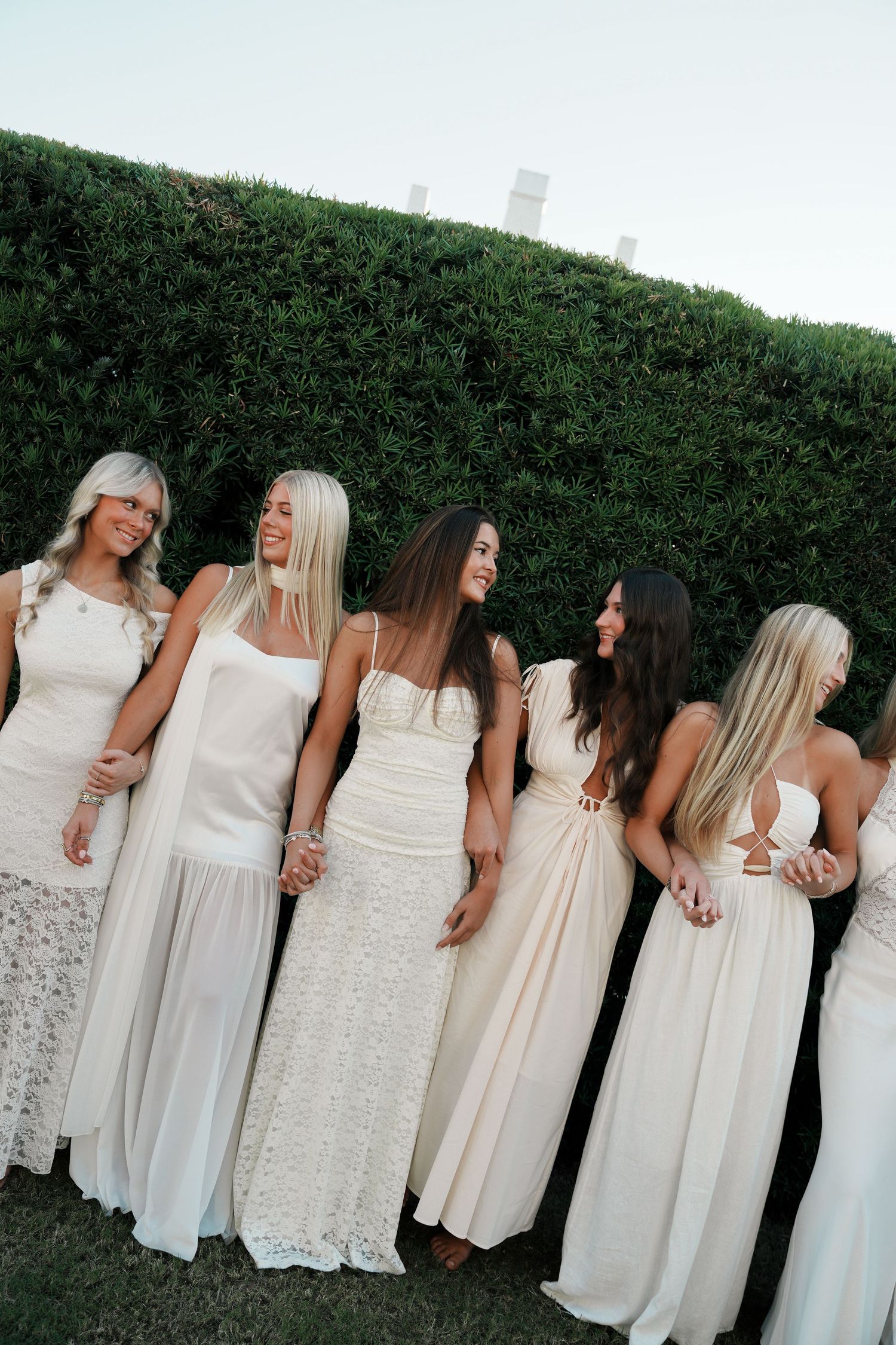 Group wearing white dresses stands in line against green hedge backdrop at outdoor event.