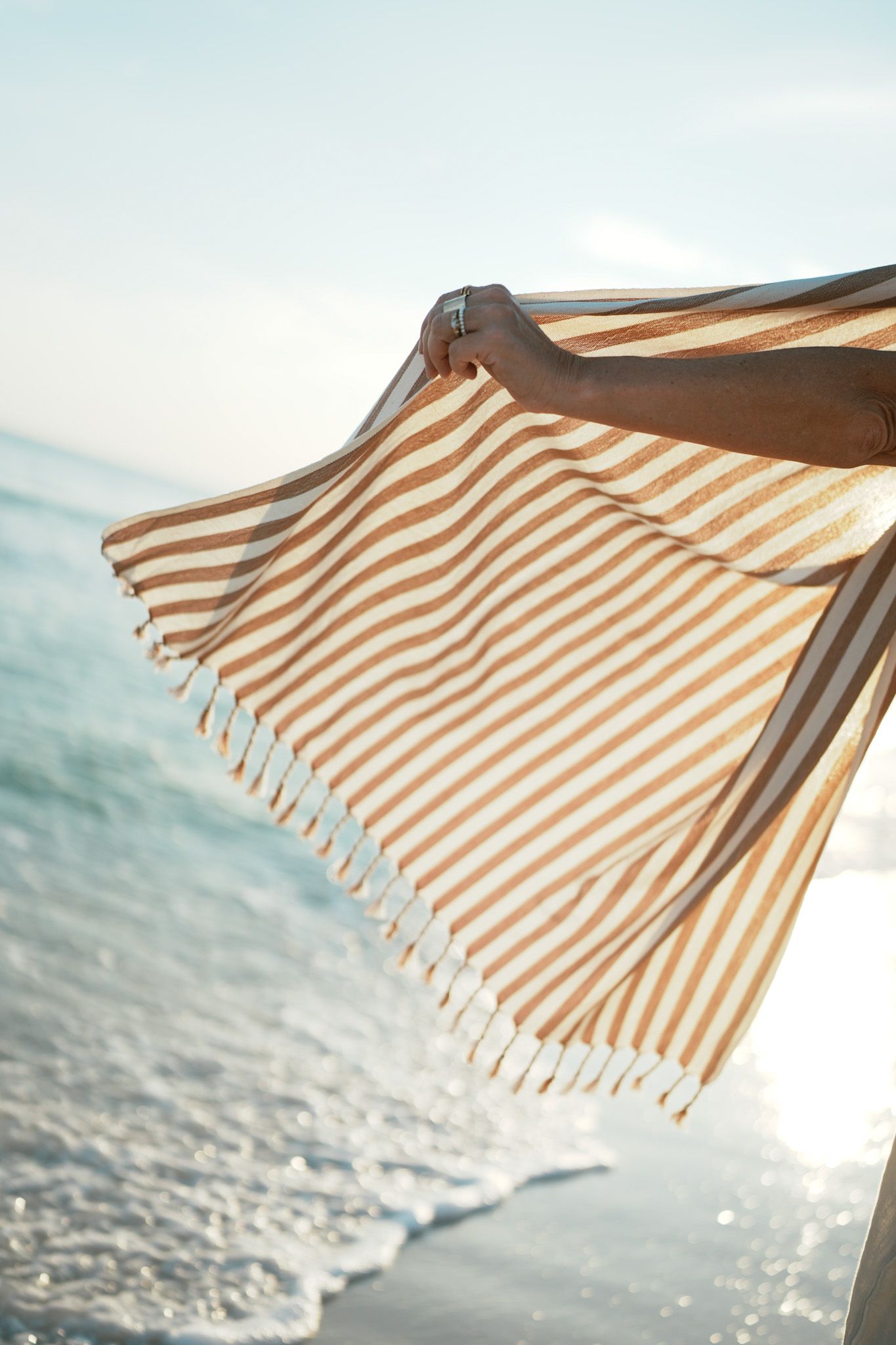 A striped beach umbrella creates geometric shadows on white sand against a bright blue ocean backdrop.