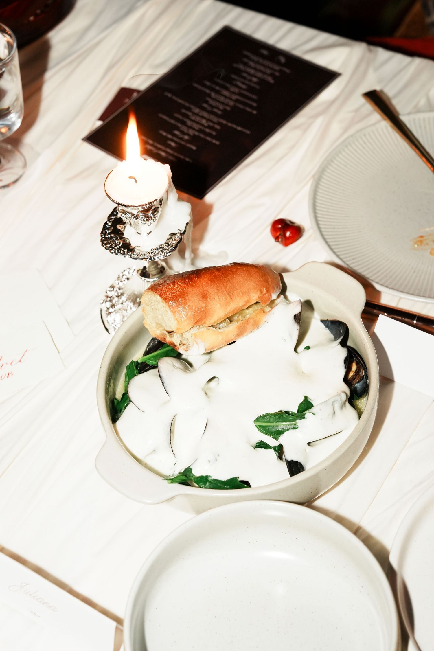 Elegant dinner table setting with bread roll salad bowl, lit candle, menu card, and white plates on linen tablecloth.