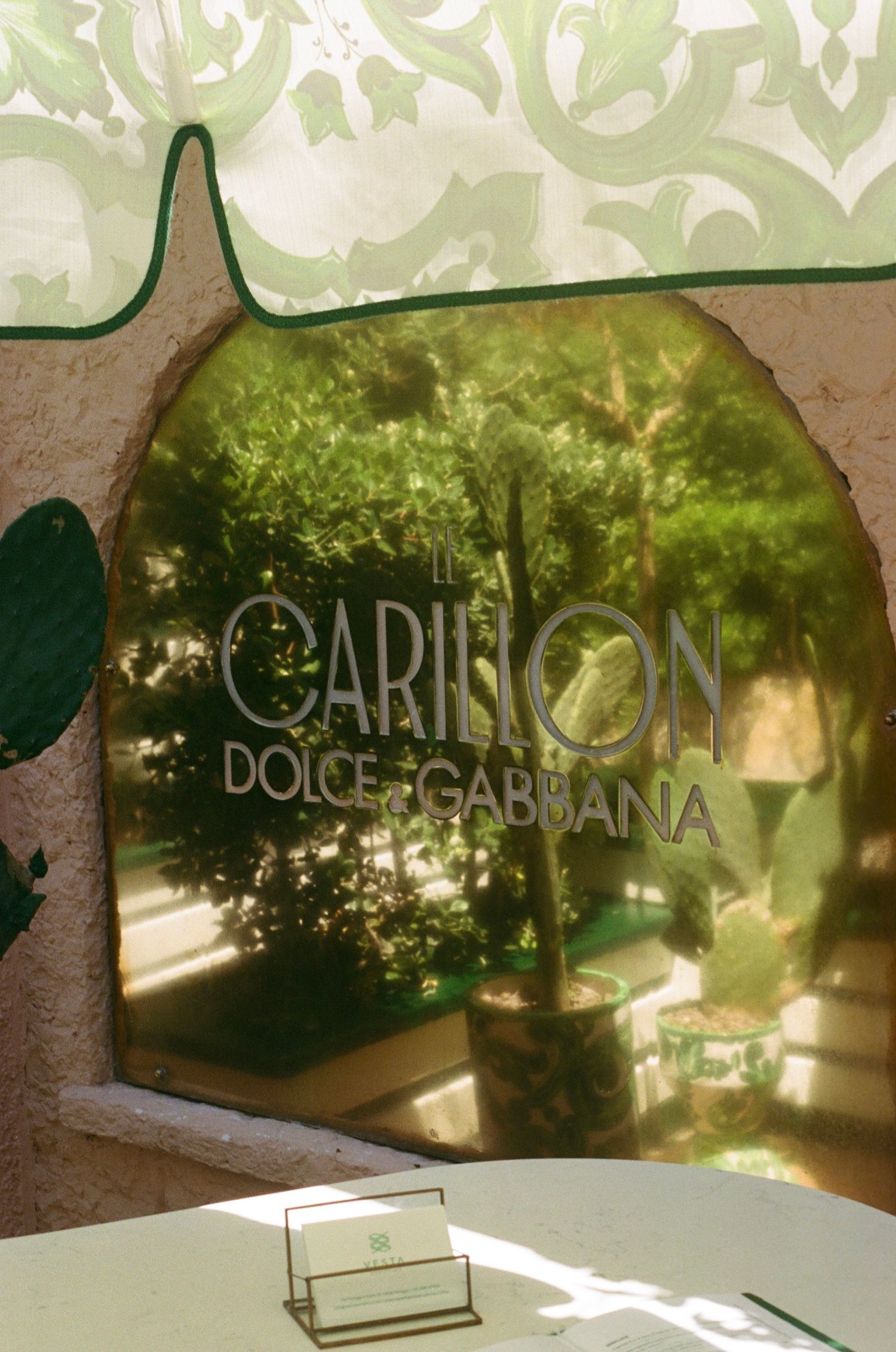 Arched mirror with Dolce & Gabbana Il Carillon branding reflecting lush green garden, set against ornate floral wallpaper.