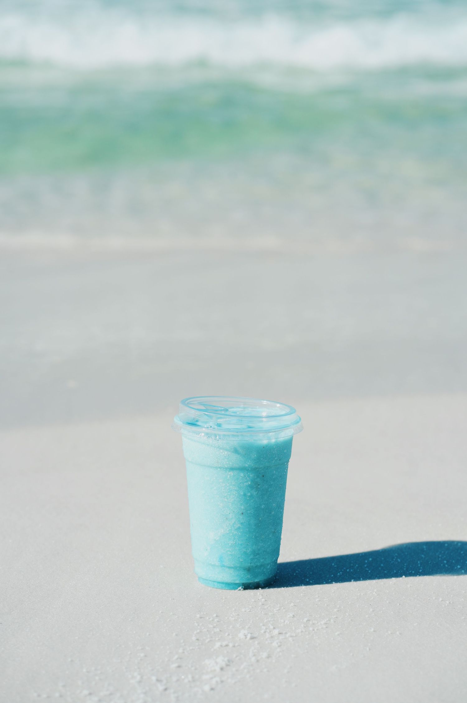 A bright blue smoothie in a clear plastic cup sits on a light sandy surface against a soft blurred background.