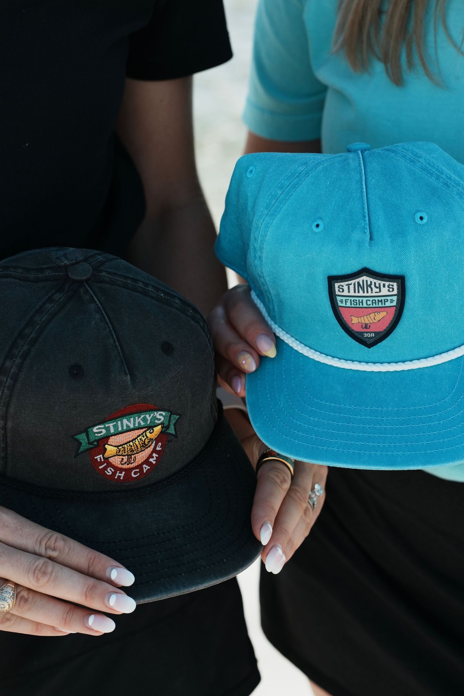 Close-up of two hands holding baseball caps with embroidered logos in black and turquoise colors.