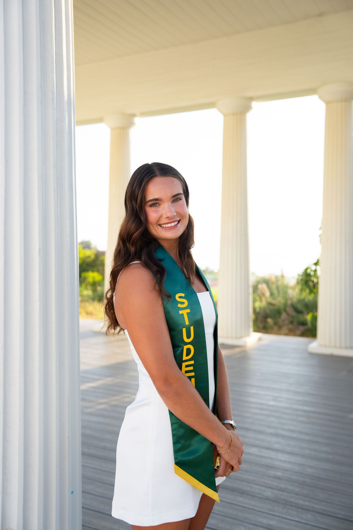 Graduate in green and gold stole poses against white columns in outdoor graduation photo session.