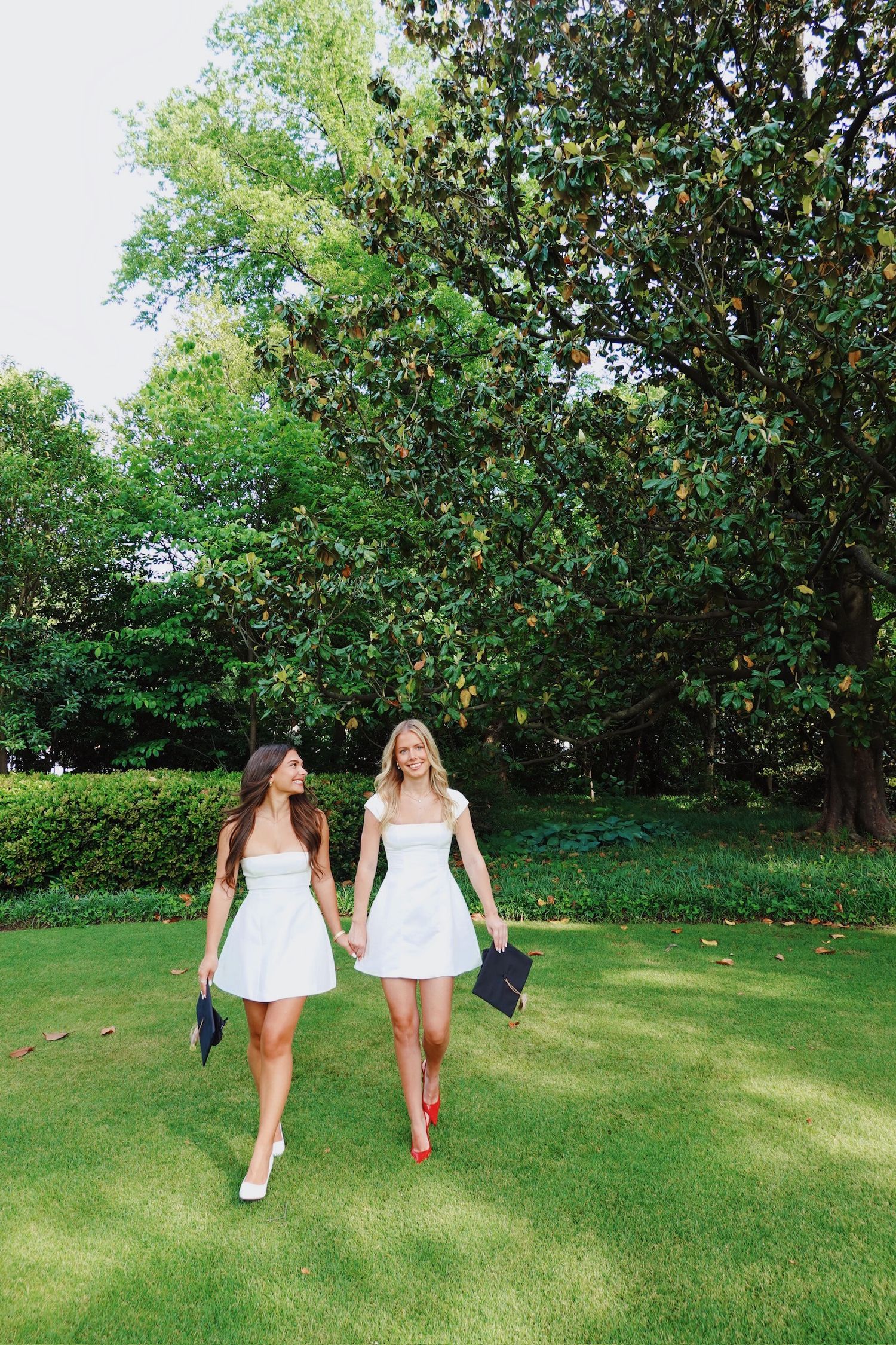 Two graduates in white dresses walking together on grass holding caps.