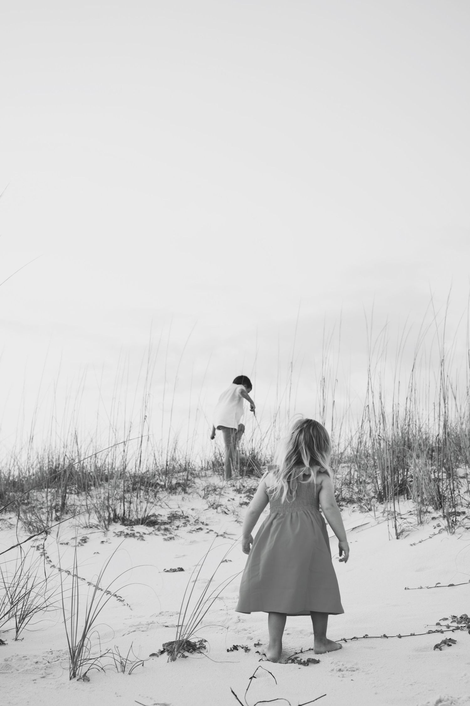 Two children walking up a sandy dune with tall grass in black and white photography.