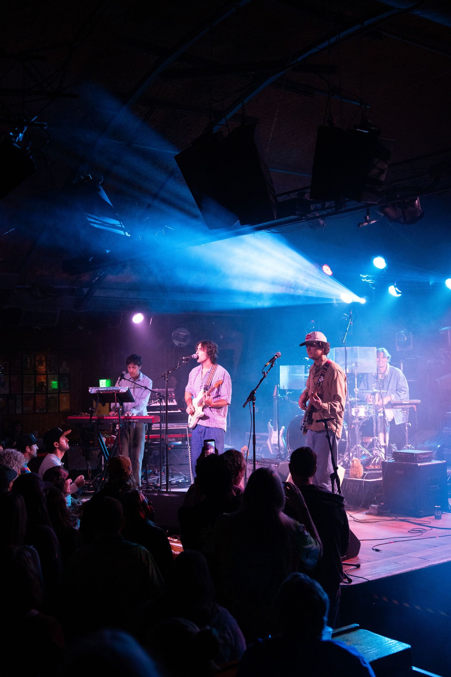 Musicians perform on stage with dramatic blue lighting and silhouetted crowd at an indoor concert venue.