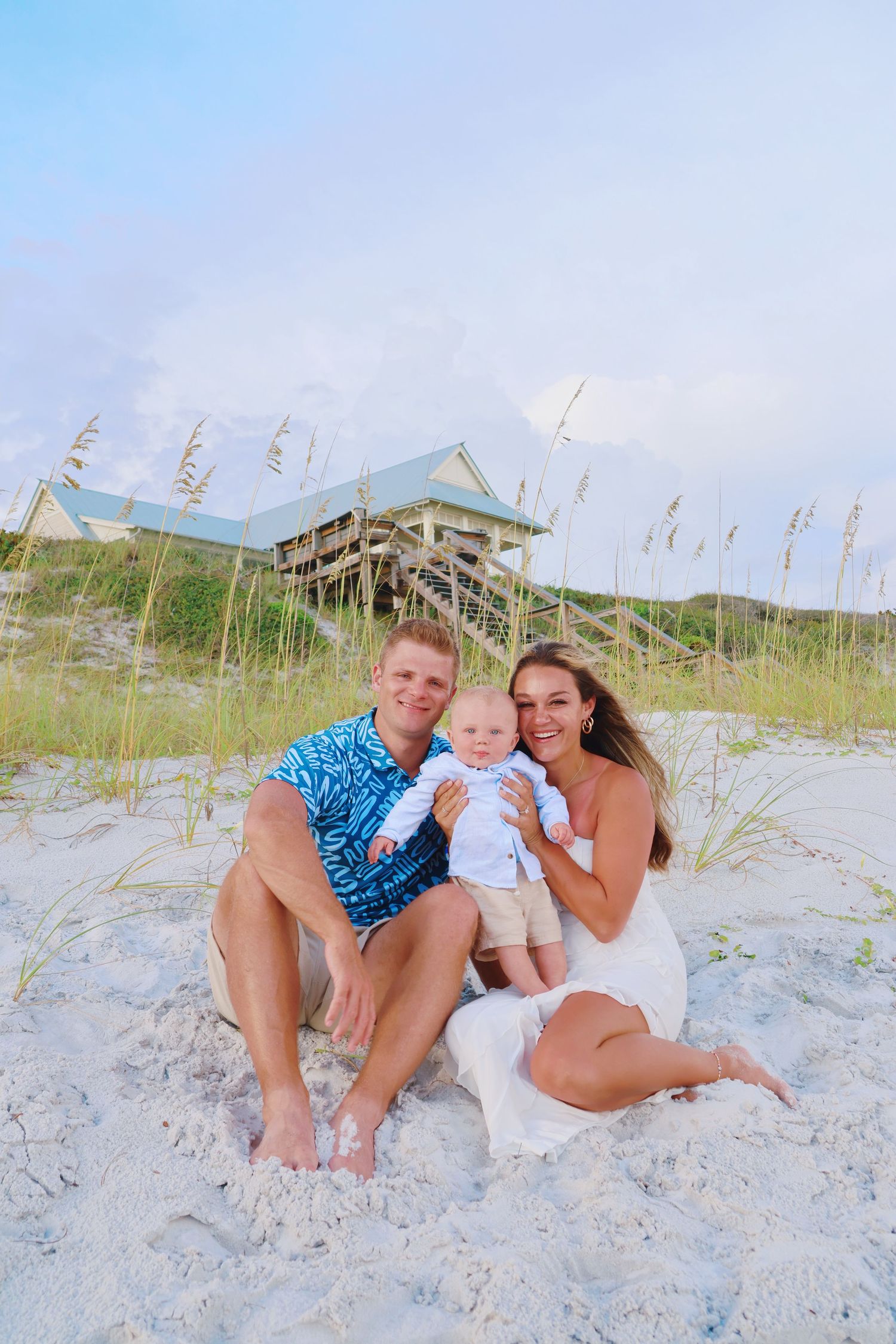 Family of three sitting together on beach sand with coastal architecture and dunes in background.