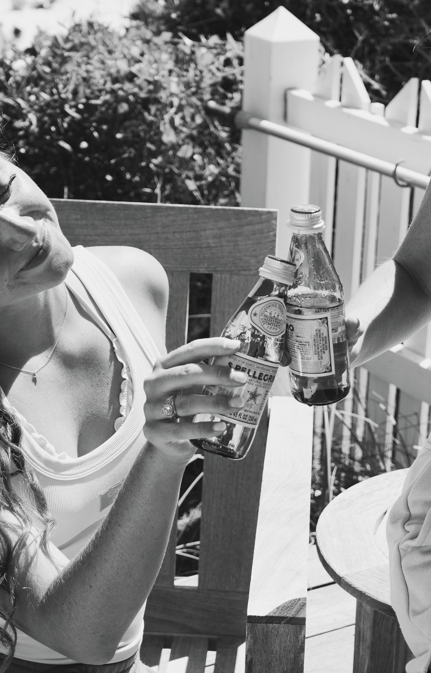 A black and white photograph of two glass bottles clinking together on a sunny porch with white fence pickets.