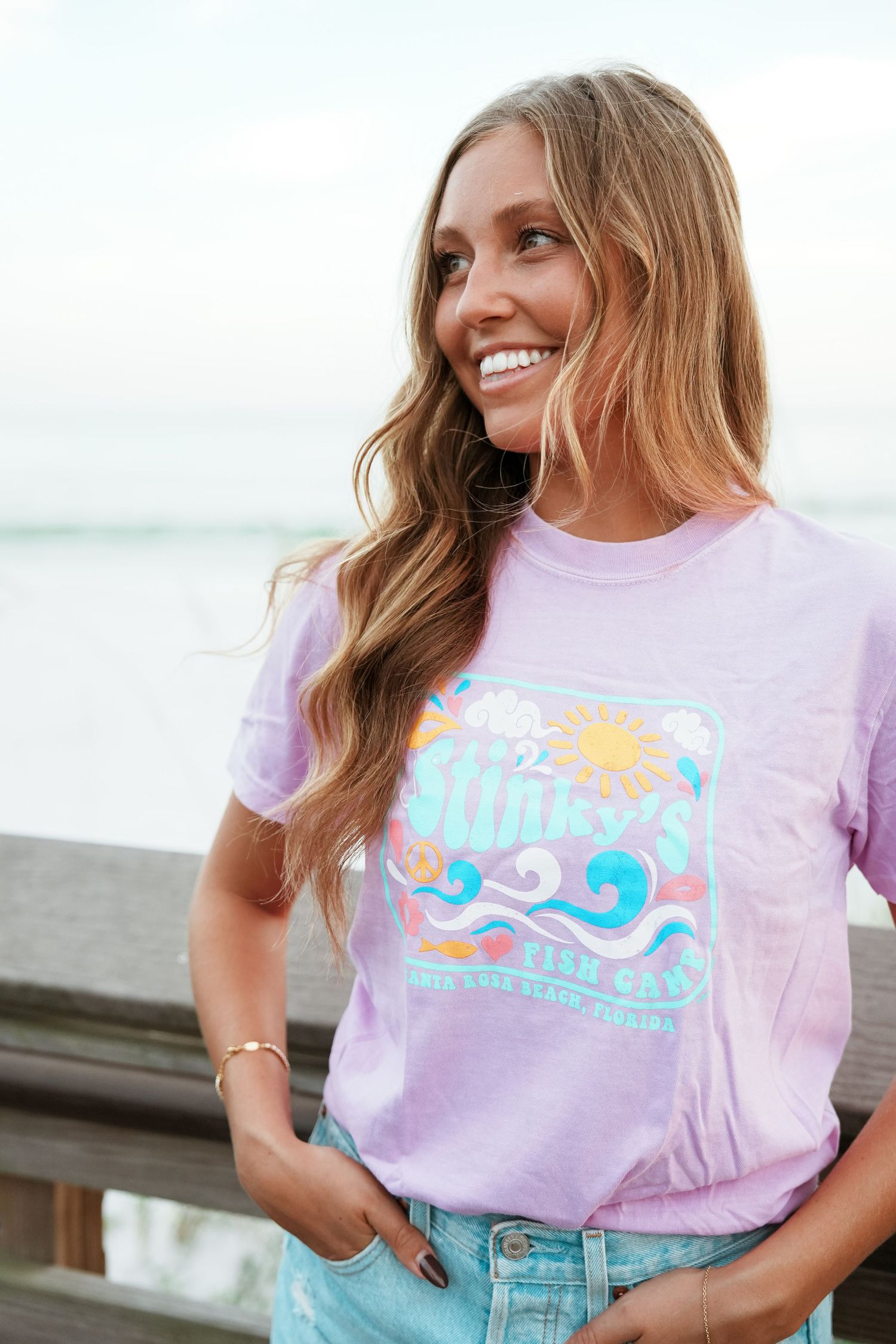 Woman wearing a pastel purple t-shirt with retro wave design and blue jeans smiles while posing outdoors.
