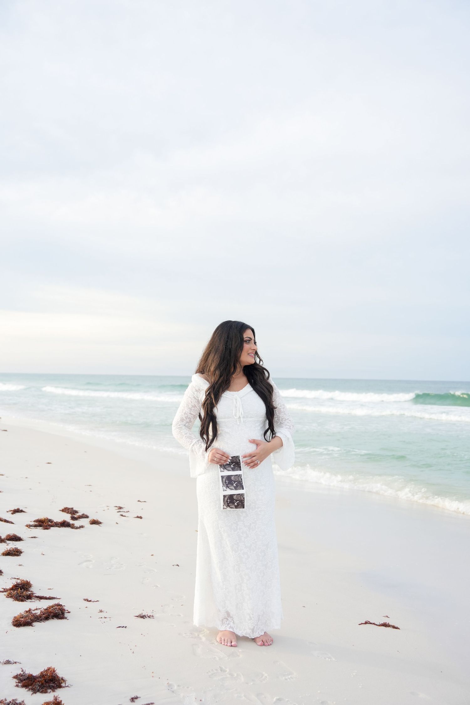 Pregnant woman in white lace gown holding ultrasound photos on a serene beach with turquoise waves.
