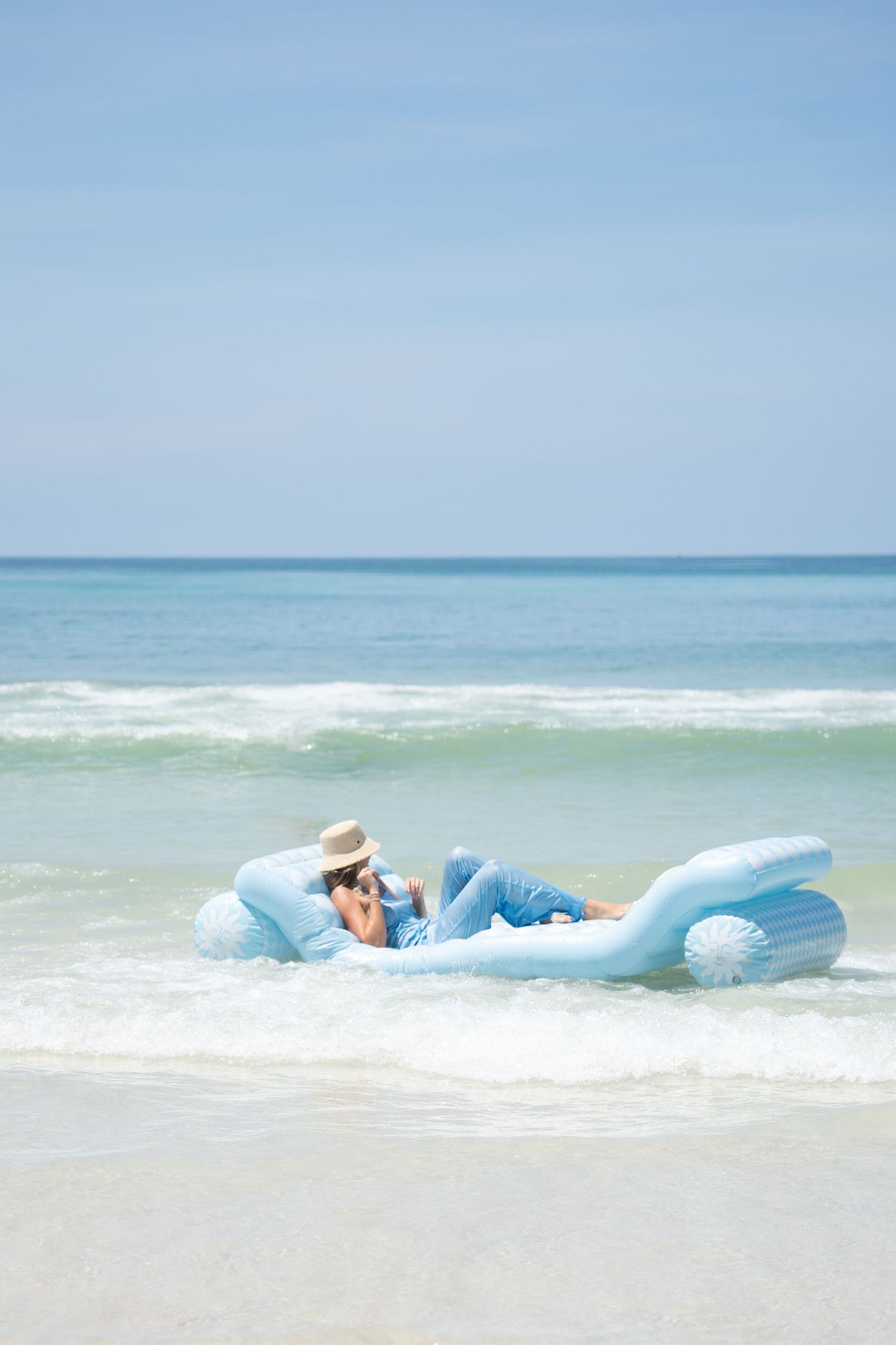 Person relaxing on a light blue inflatable pool float in ocean waves at a sunny tropical beach.