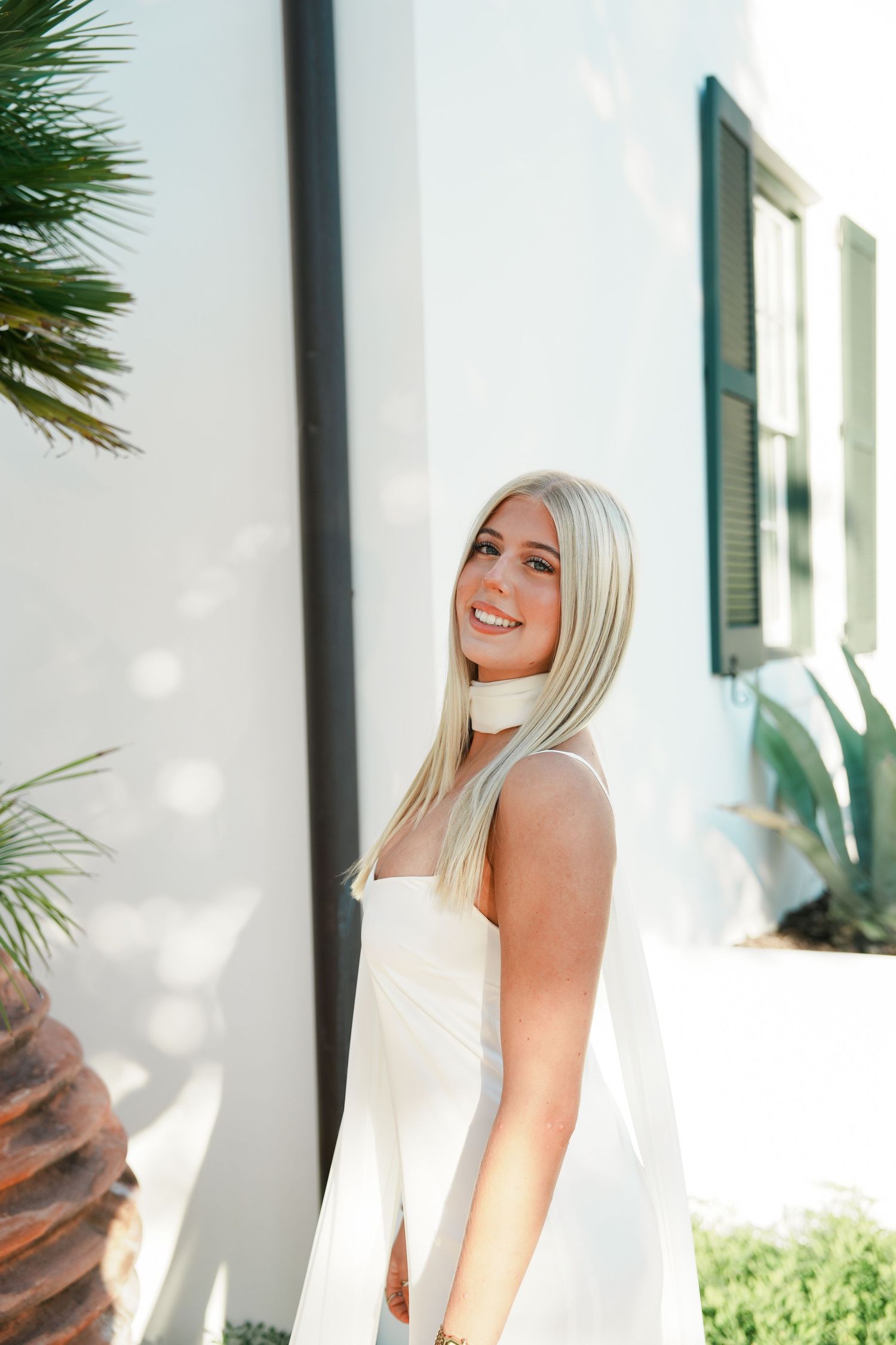 Someone in a white summer dress stands against a white wall with palm fronds and window shutters visible in the background.