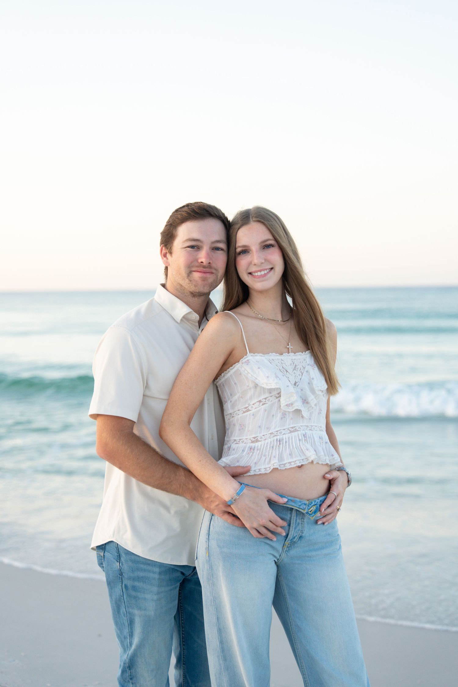 A couple poses together on a sandy beach, smiling, wearing white tops and jeans with ocean waves behind them.