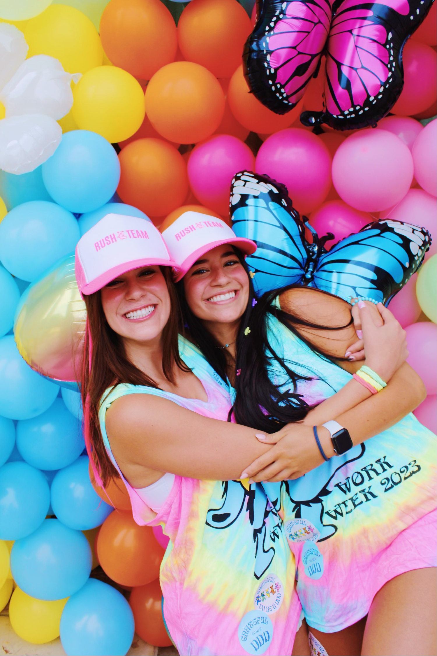 Group wearing pink shirts and hats poses against colorful balloon backdrop with decorative butterflies.