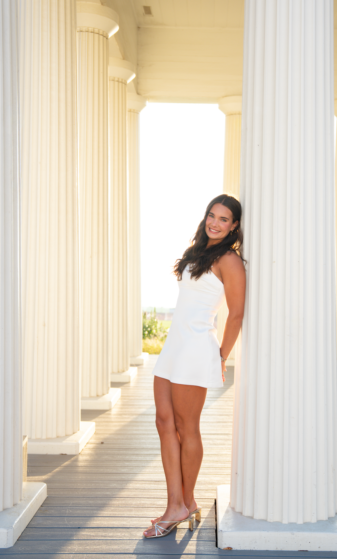 Woman in white dress poses playfully between classical white columns in an architectural setting.