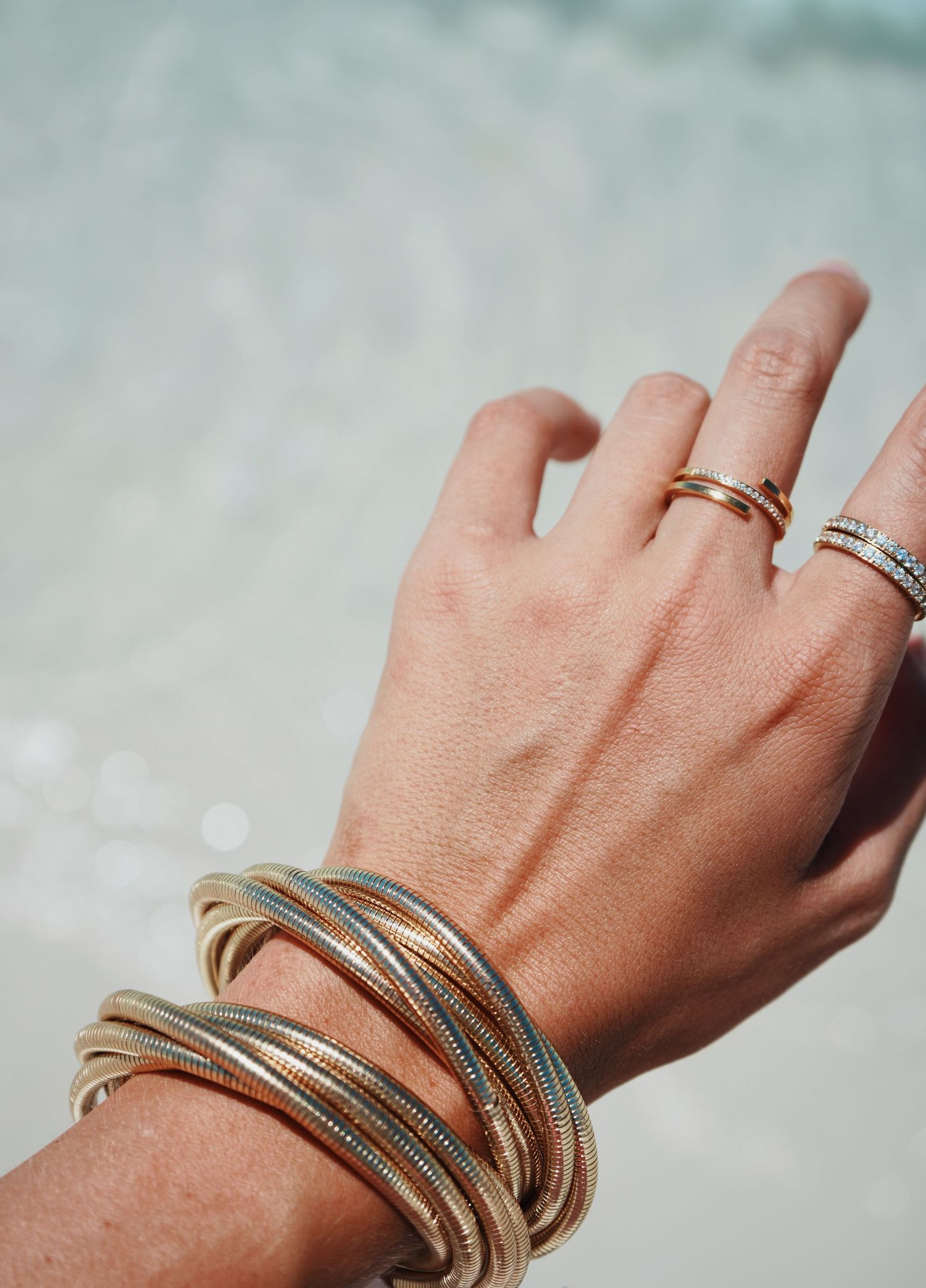 Hand wearing layered metallic bangle bracelets and stacked rings in silver tones against blurred light background.