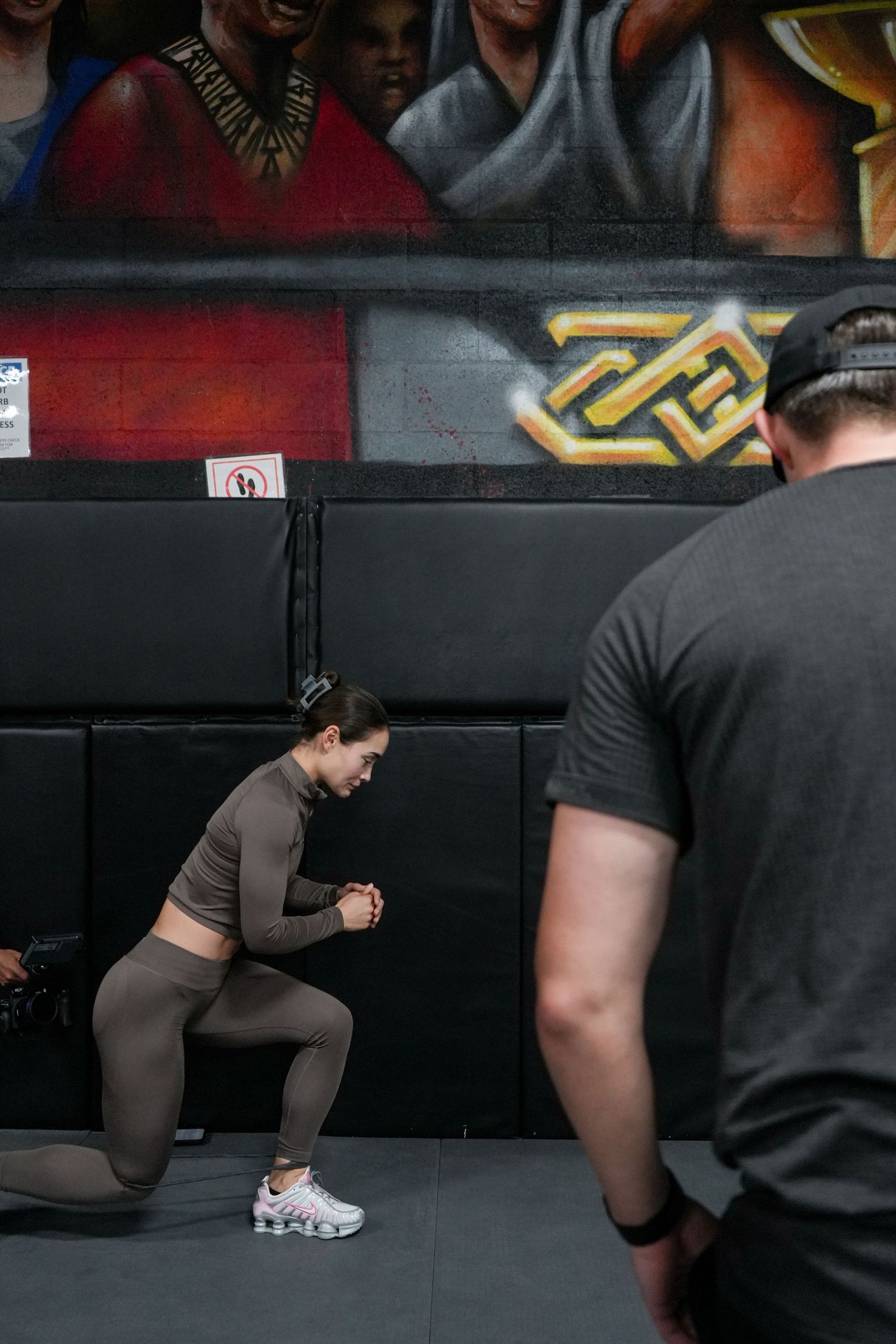 Person in athletic wear performs a lunging exercise in a gym setting with dark walls and graffiti.
