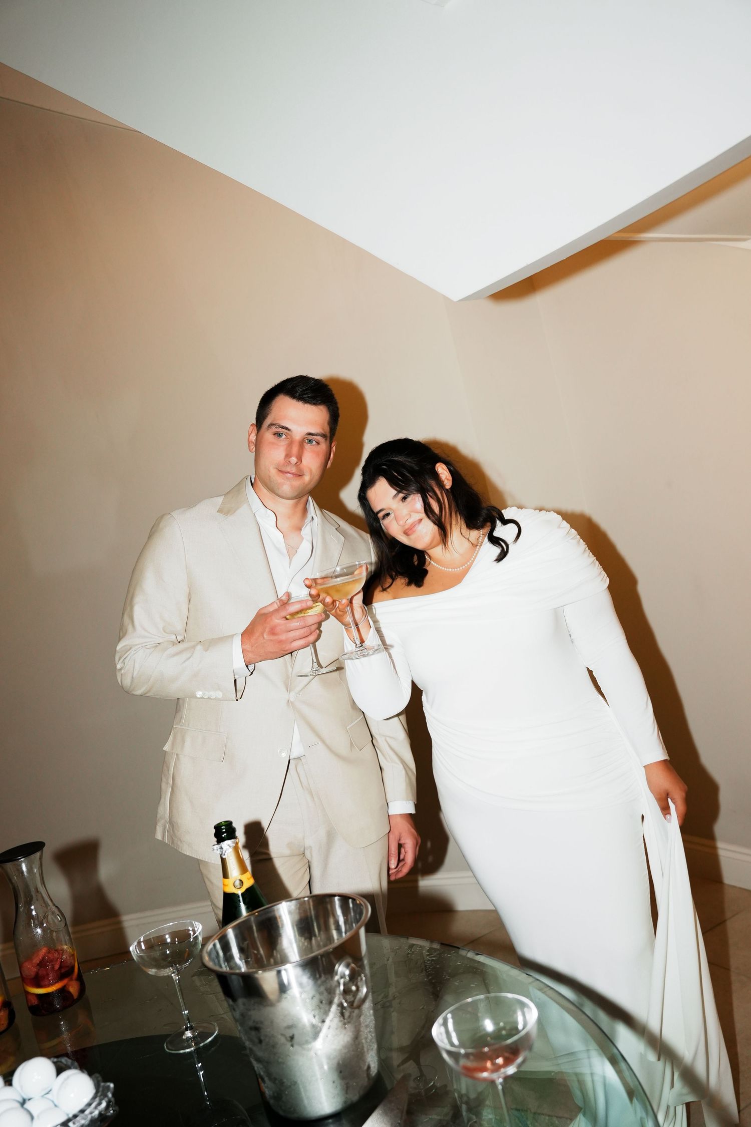 Two people in white outfits stand near a table with drinks and an ice bucket at what appears to be a party setting.