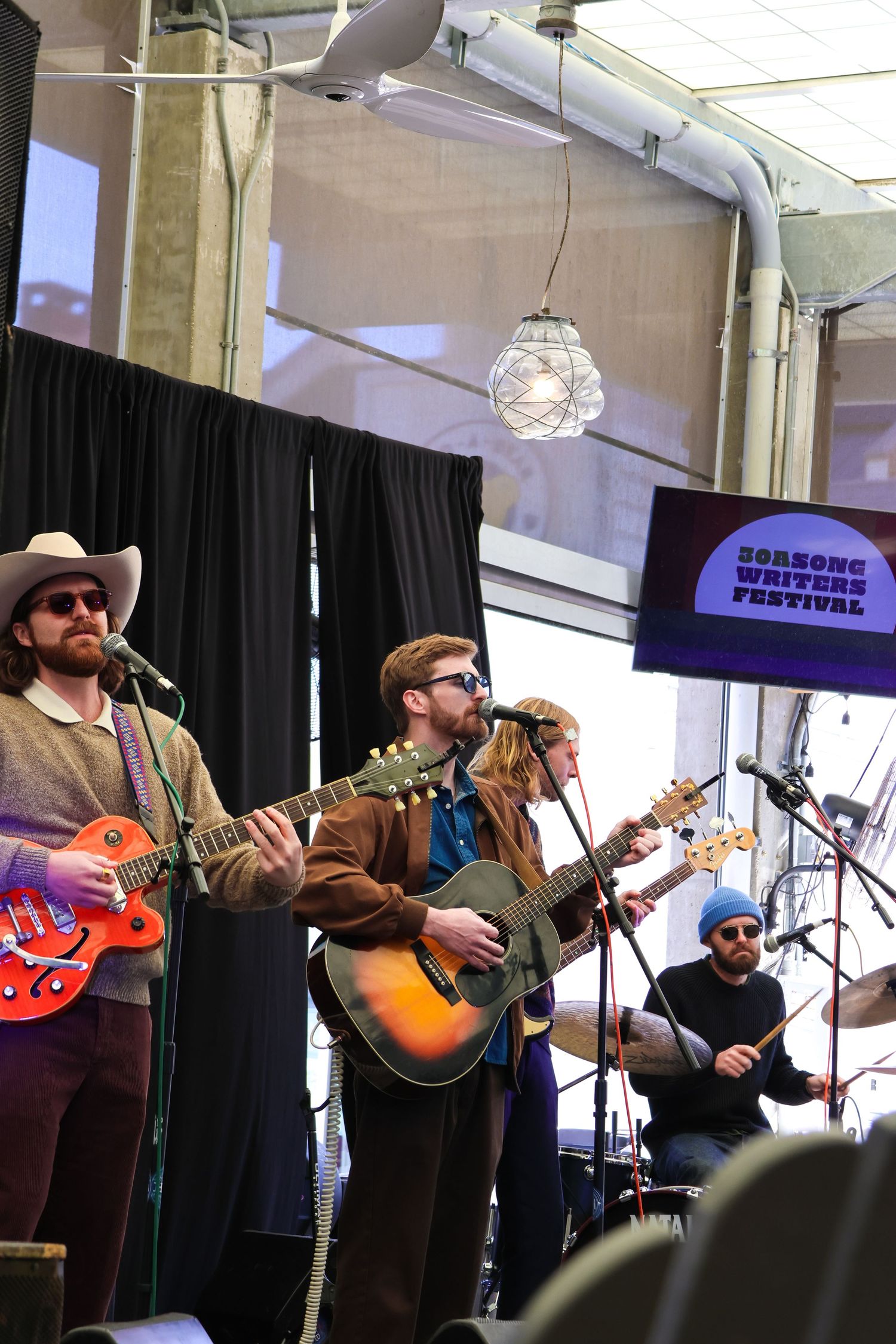 Folk band performing with guitars under festival lighting with banner visible in background.