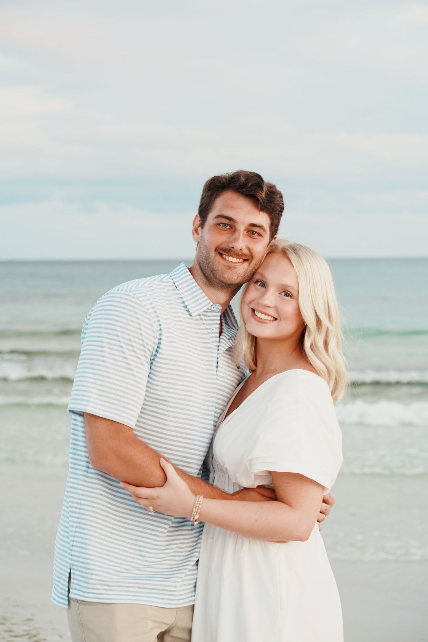 Happy couple embraces on beach at sunset wearing white and striped outfits against ocean backdrop.