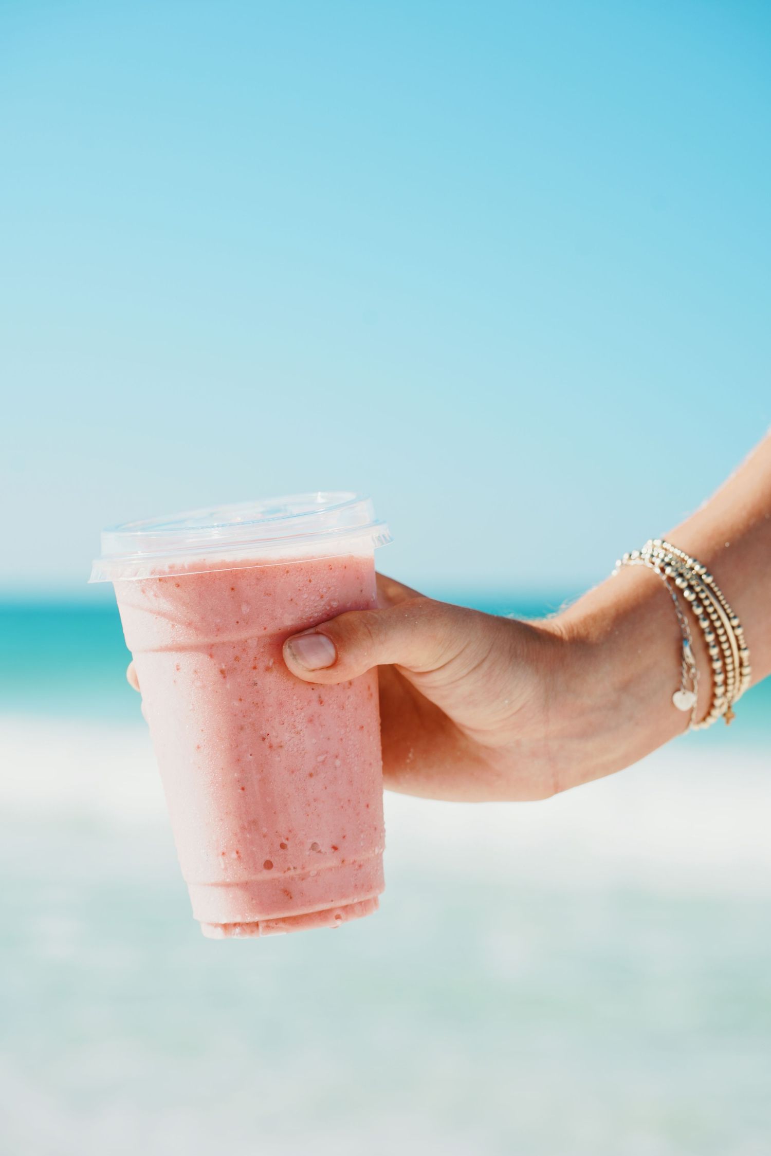 Hand holding a pink strawberry smoothie against a bright turquoise ocean and beach background with stacked bracelets.