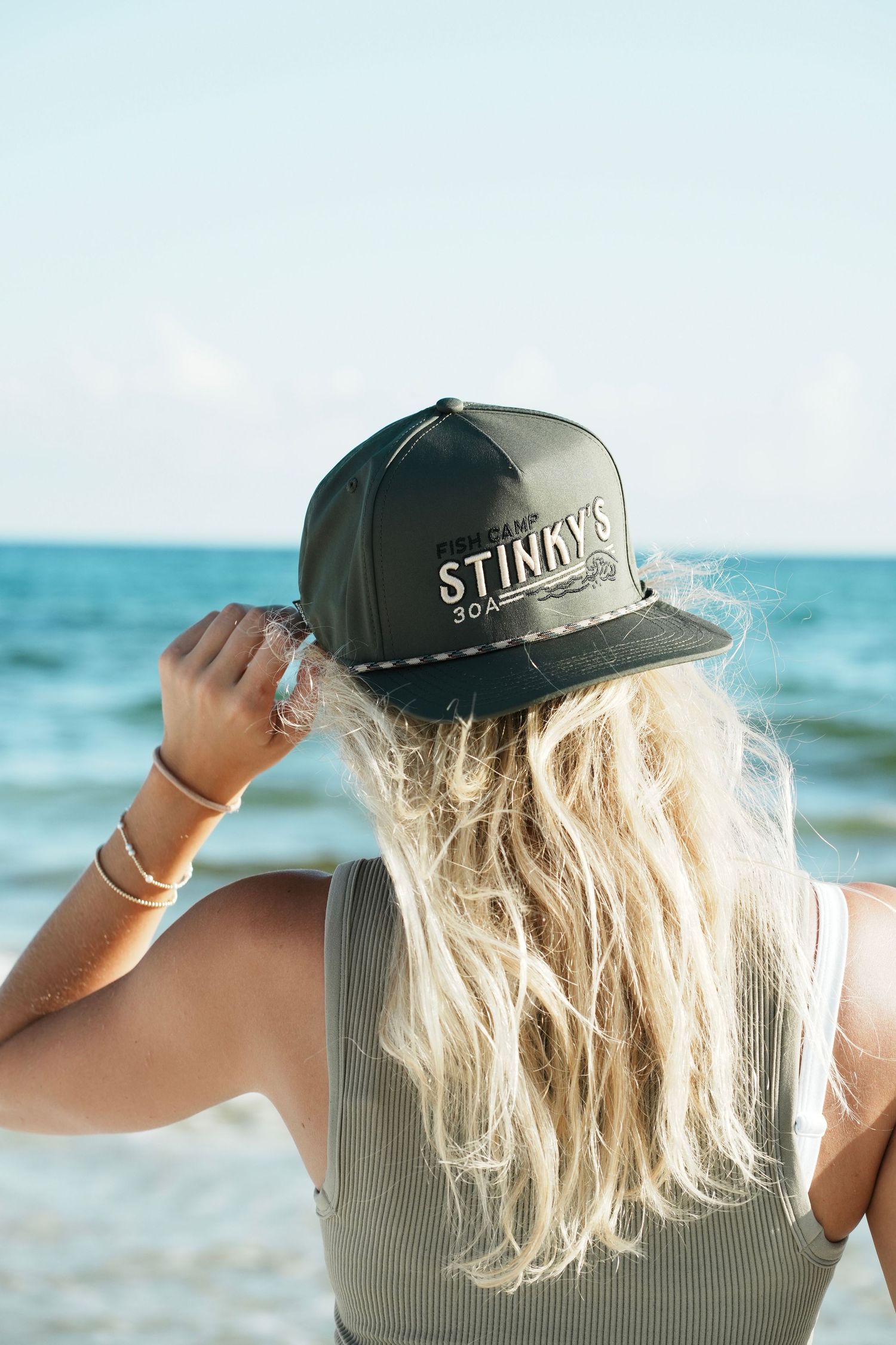 A person wearing a black baseball cap with blonde hair looks out towards turquoise ocean waters on a sunny day.