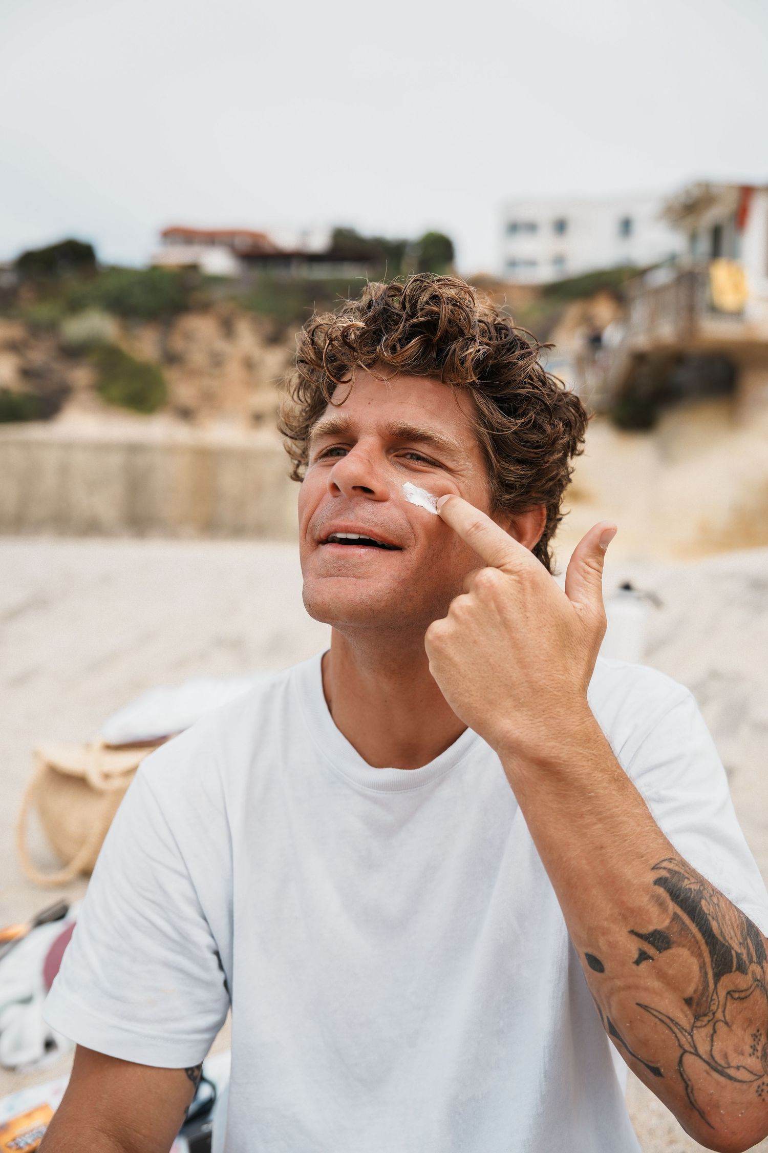 Person in white t-shirt applying sunscreen to face at beach setting.