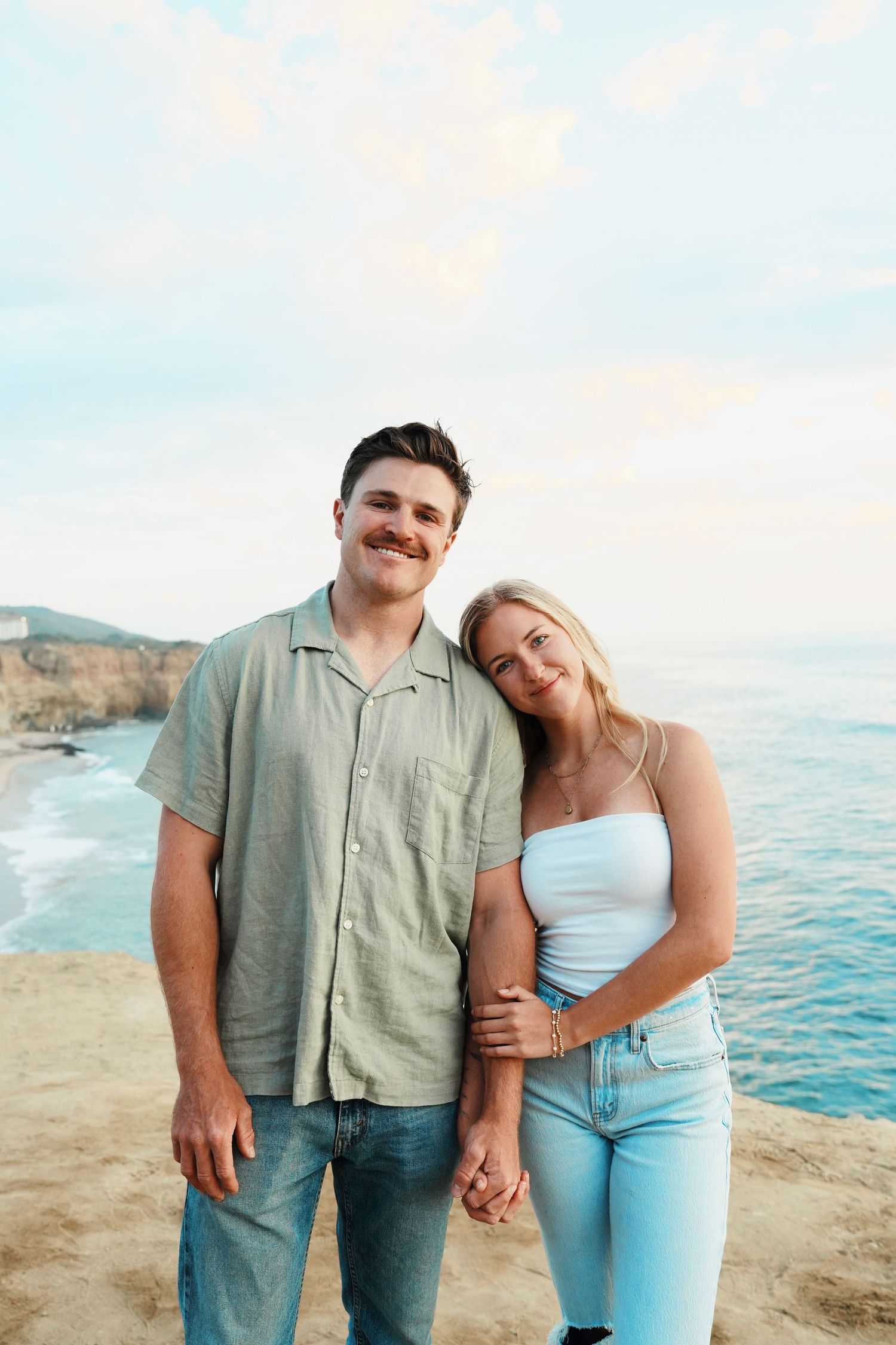 A couple stands together on a coastal cliff overlooking the ocean at sunset.
