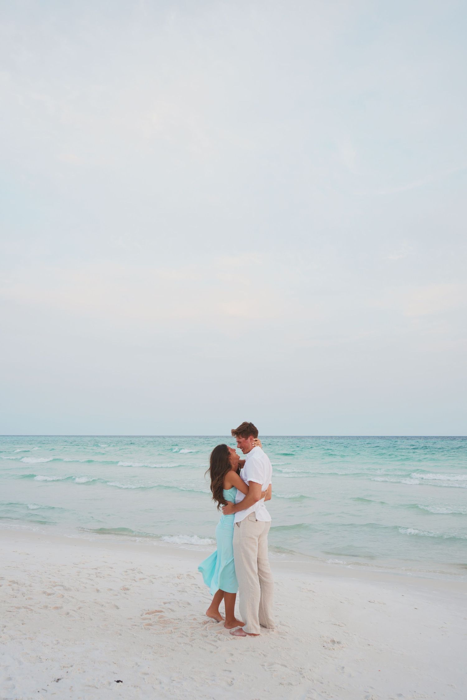 A couple in turquoise and white outfits share a romantic embrace on a tranquil beach with crystal blue waters.