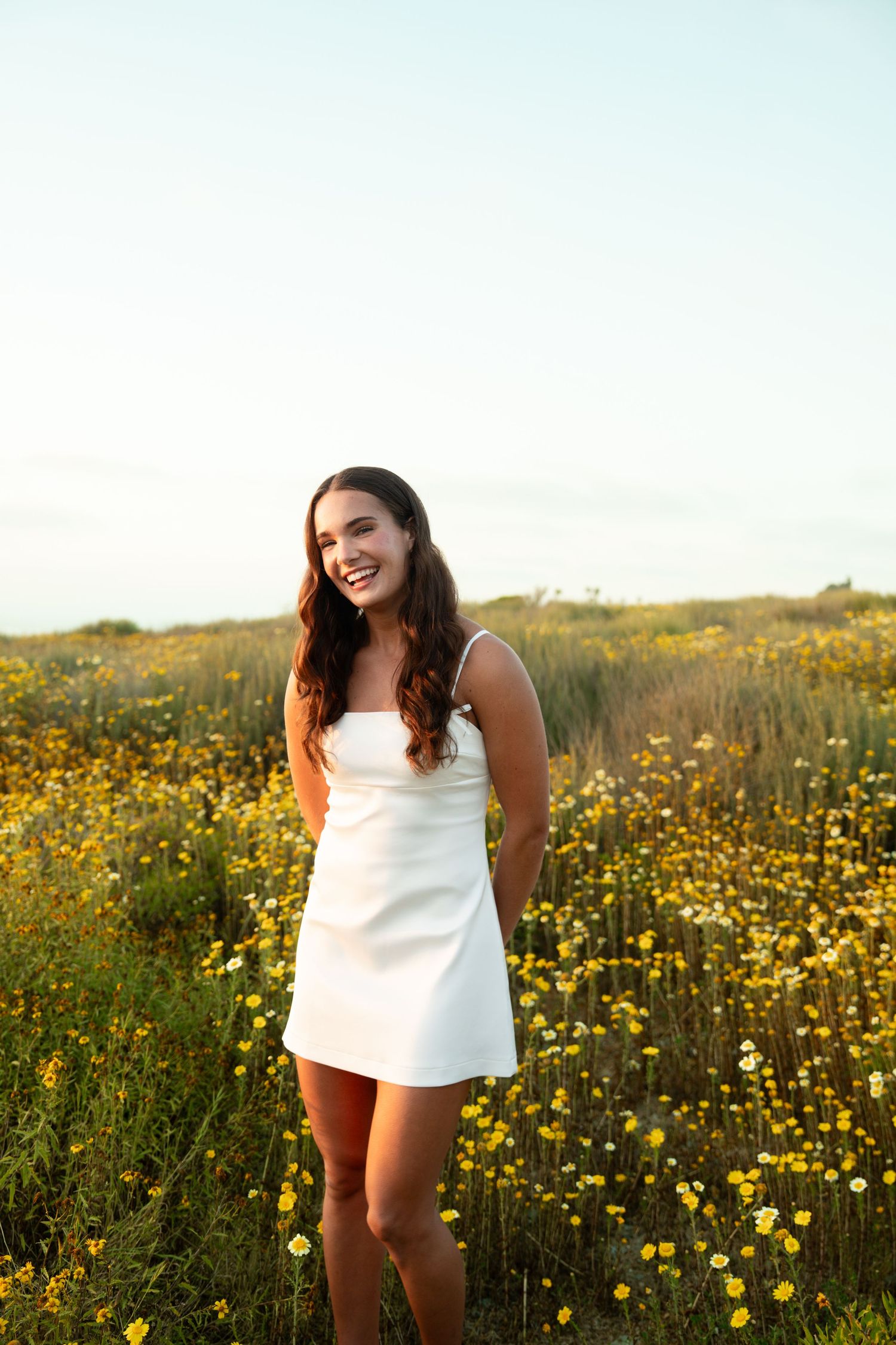 A woman in a white top stands joyfully among a vibrant field of yellow wildflowers on a sunny day.