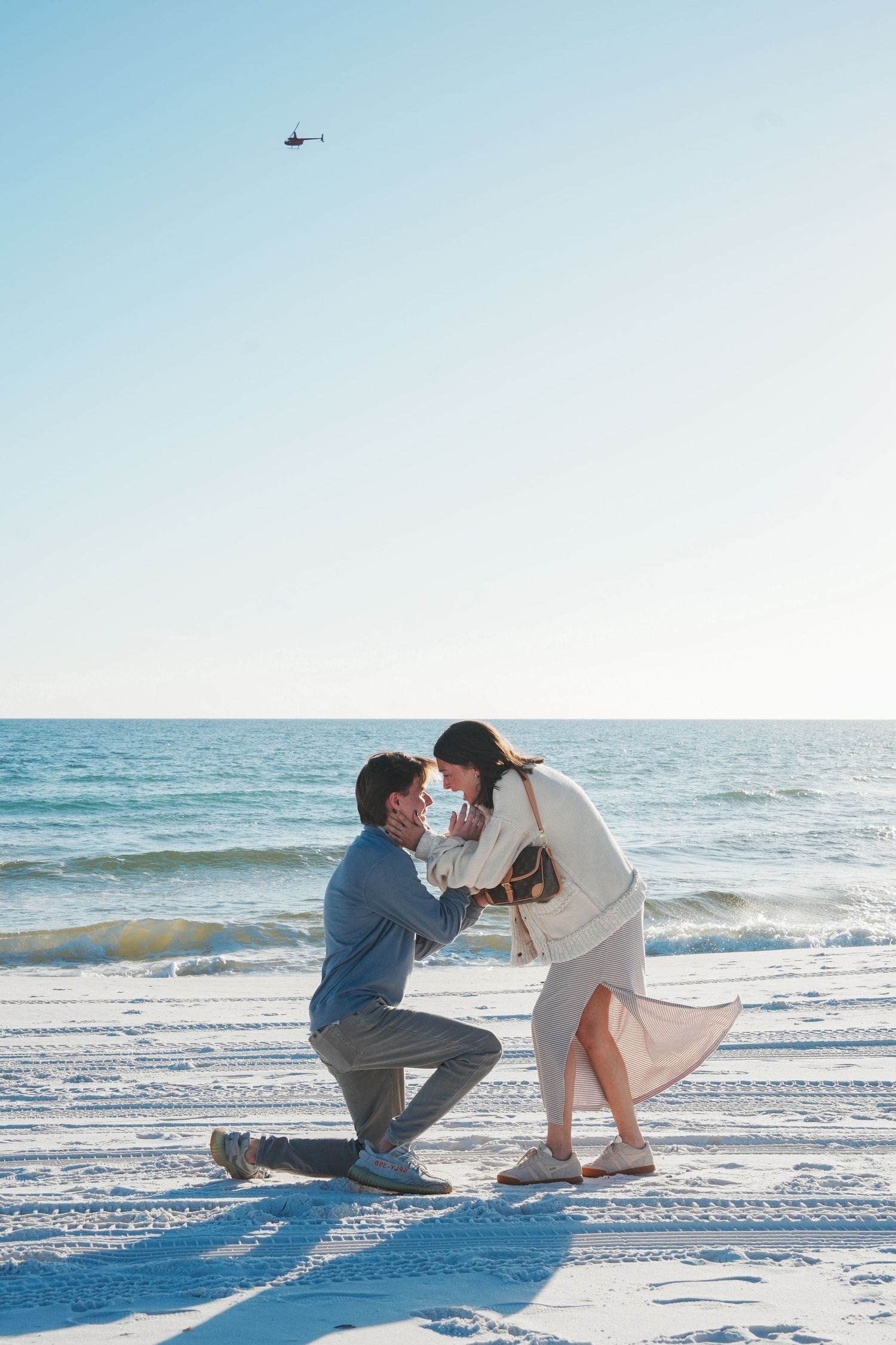 A romantic beach proposal scene with a couple on white sand as waves roll in and a helicopter flies overhead in the blue sky.