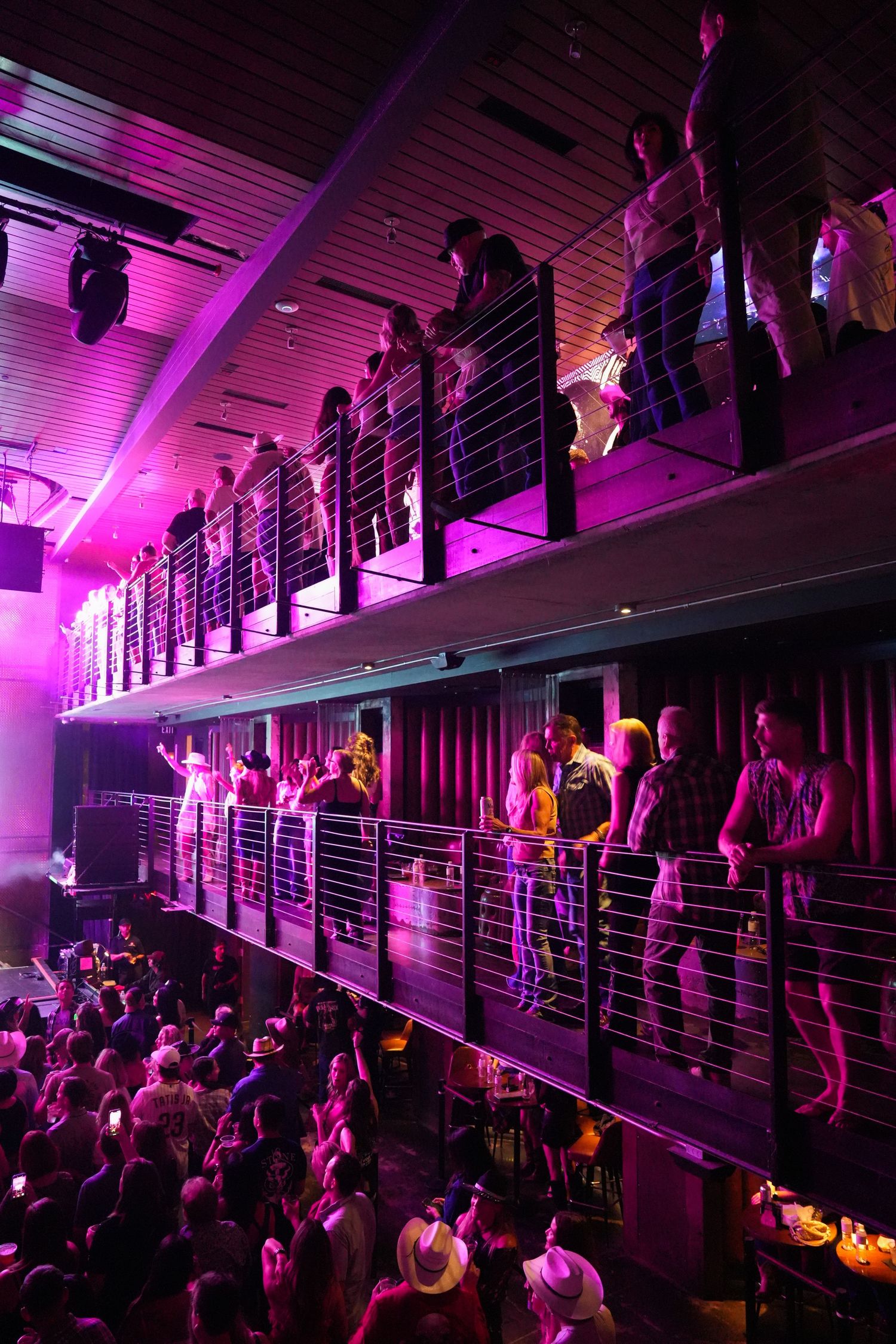 Silhouettes of concert attendees standing on multiple levels bathed in vibrant purple lighting at a nightclub venue.