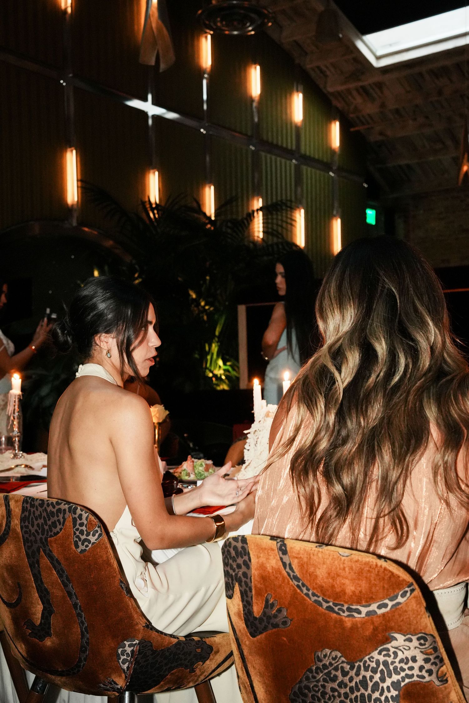 Two women sitting at a candlelit dinner table with leopard print chairs in a warmly lit elegant restaurant.