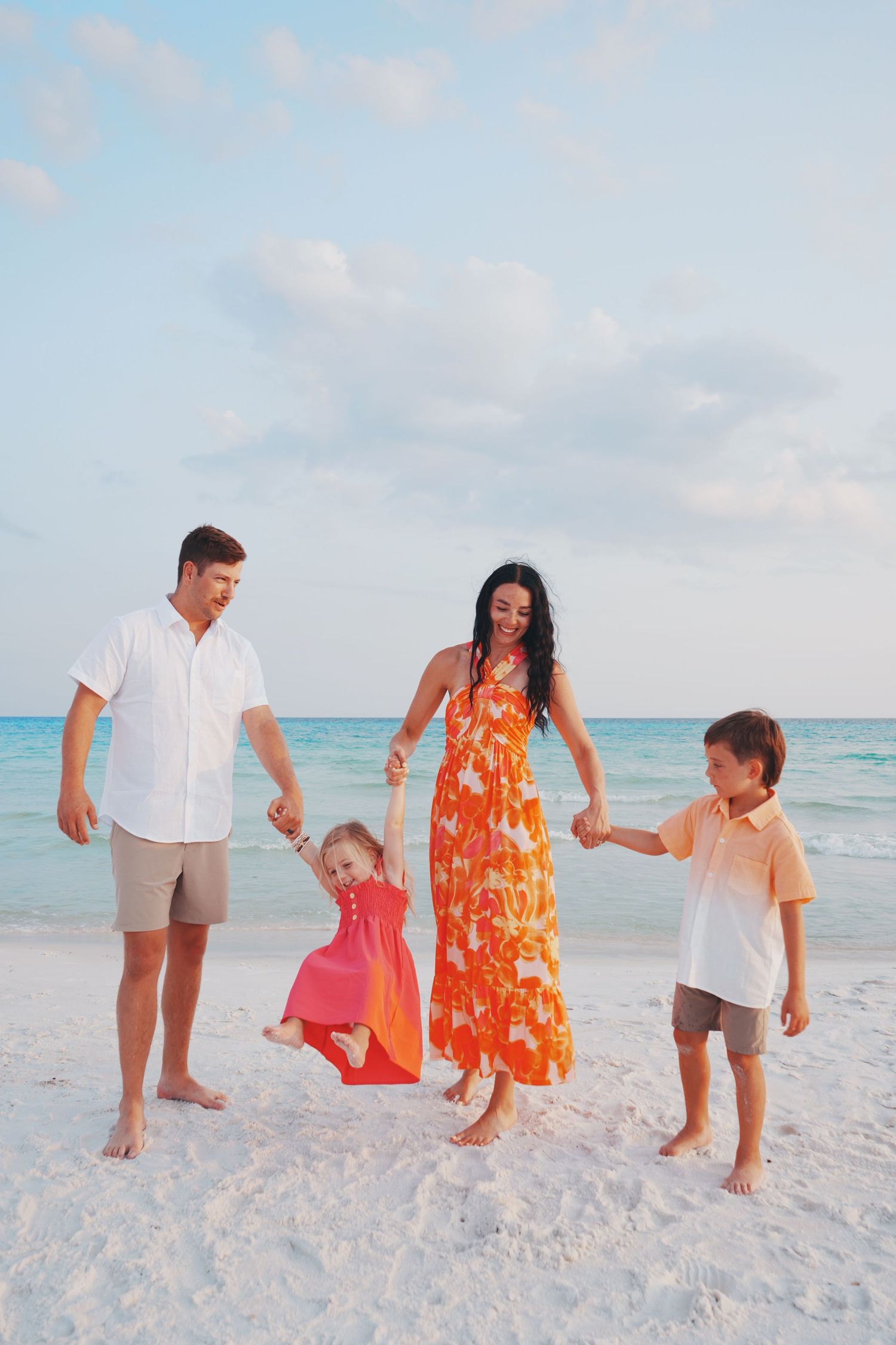 Family enjoys playful moment on white sandy beach with turquoise ocean waters and soft pastel sky in background.