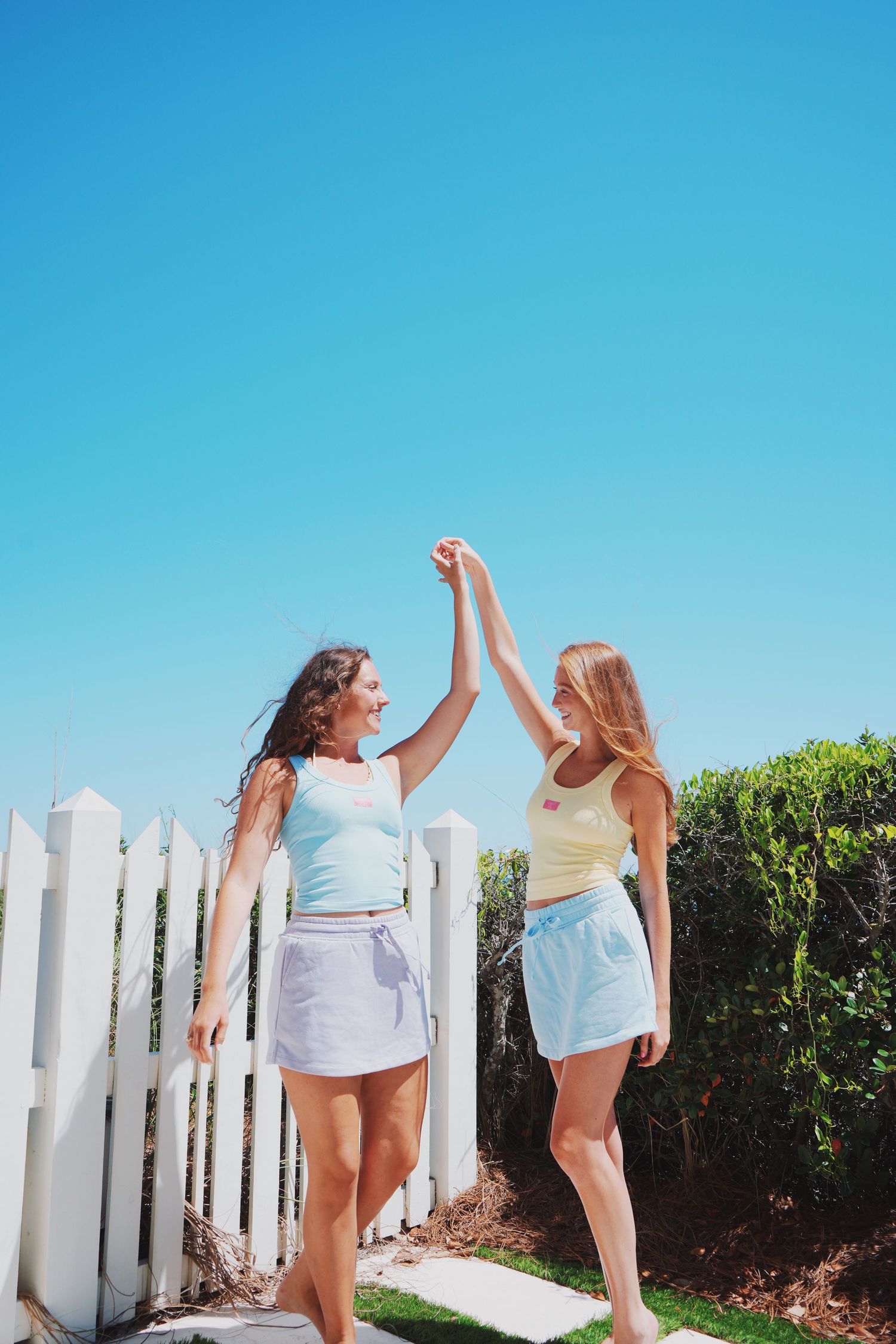 Two friends in summer outfits high-five in front of white picket fence by the beach.