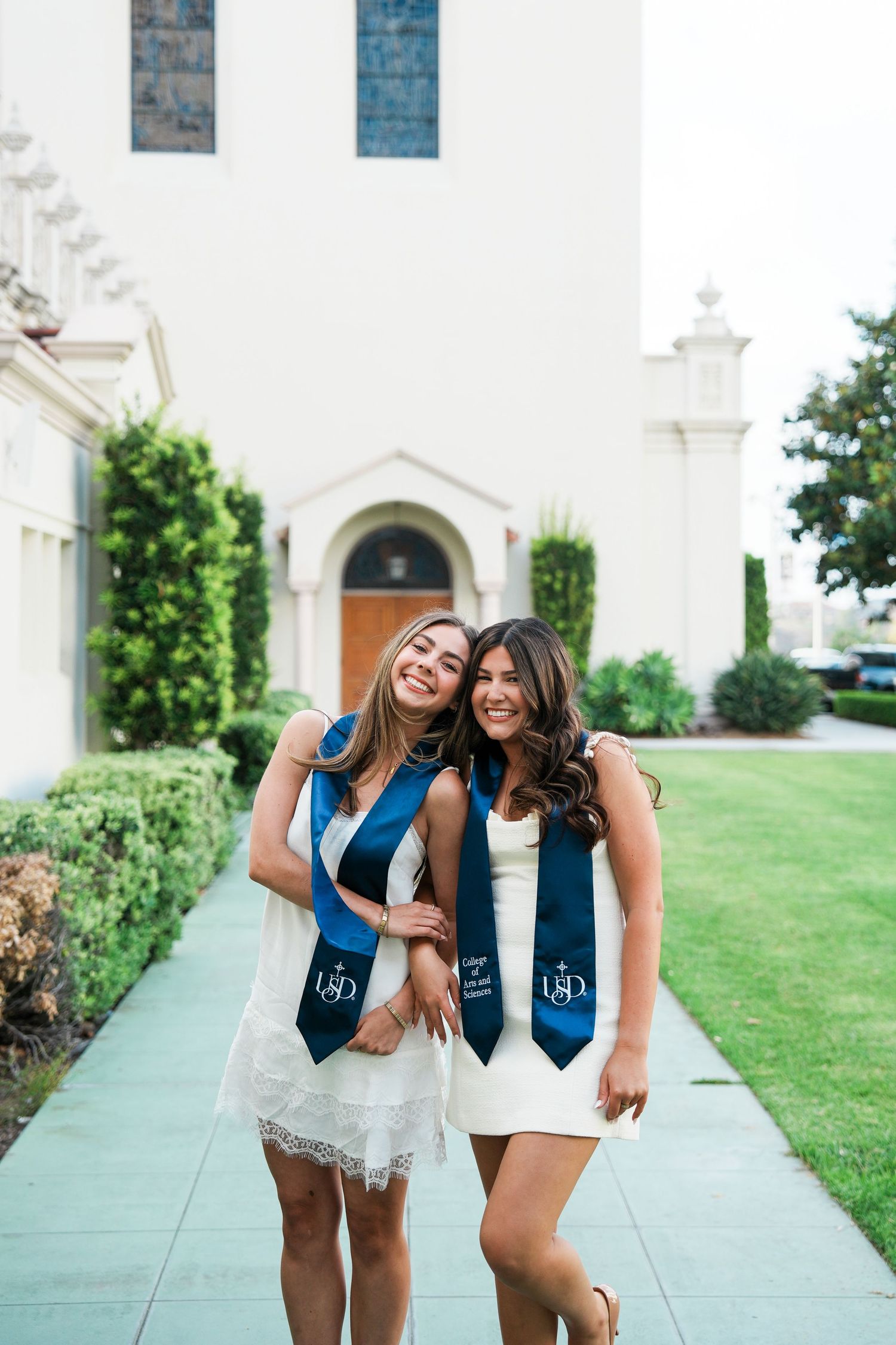 Two smiling female USD graduates wearing navy blue stoles pose together on a campus walkway in white dresses.