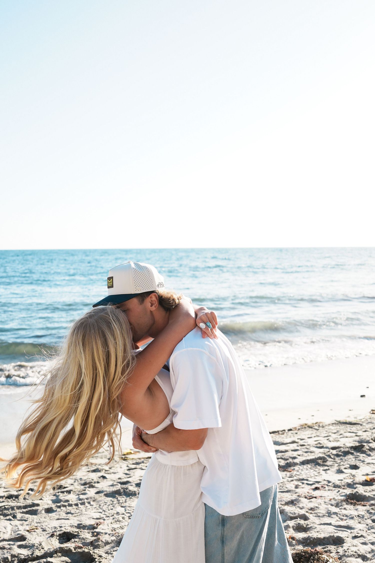 Couple kissing on a sunny beach, woman in white dress with blonde hair, man in white shirt and cap, ocean in background.